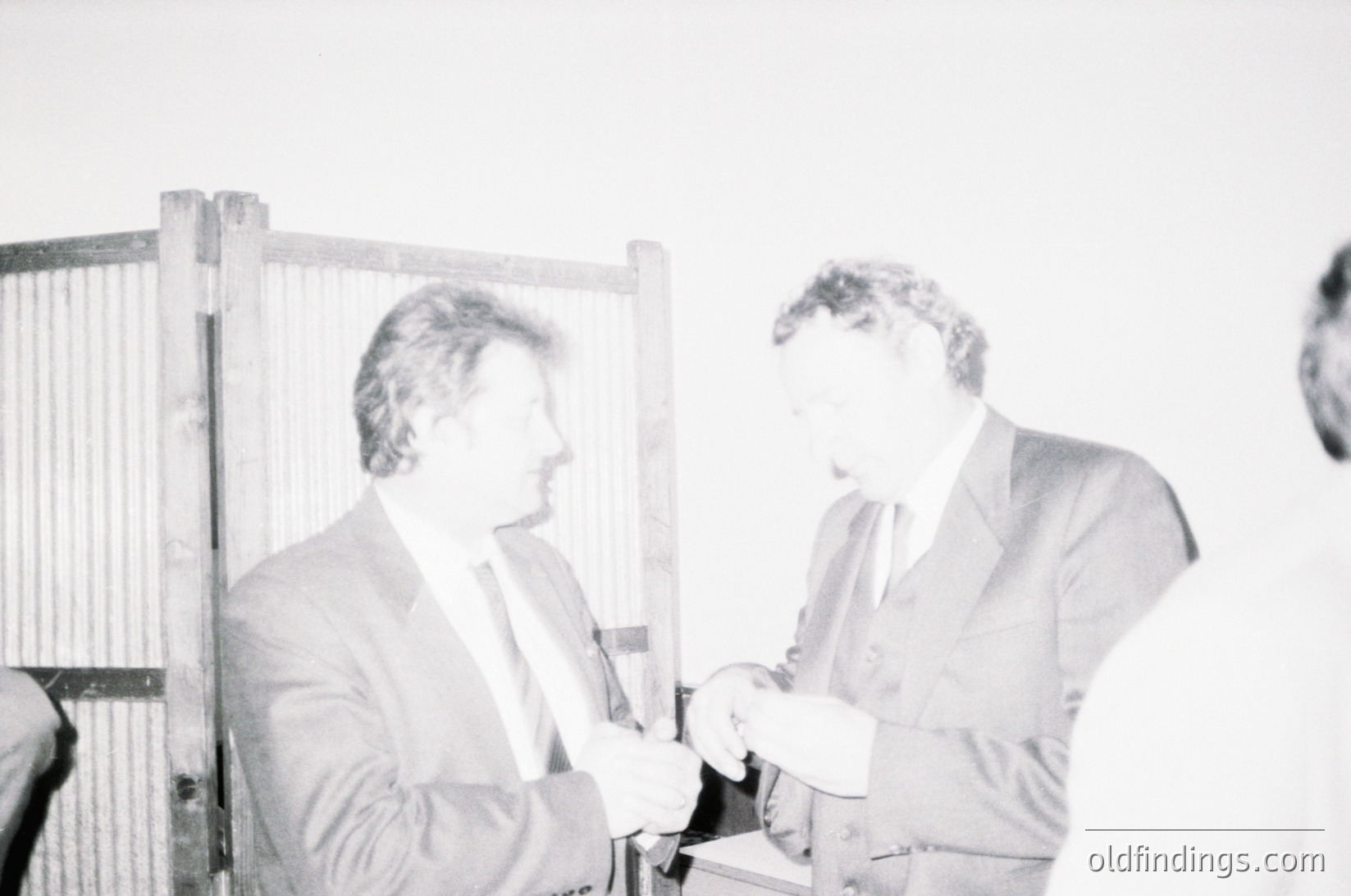 Two men in formal attire exchange a small object at an indoor event, likely mid-1970s–1980s based on clothing styles. The blurred background suggests a formal setting, possibly a conference or ceremony. The wooden paneling hints at institutional or governmental architecture.