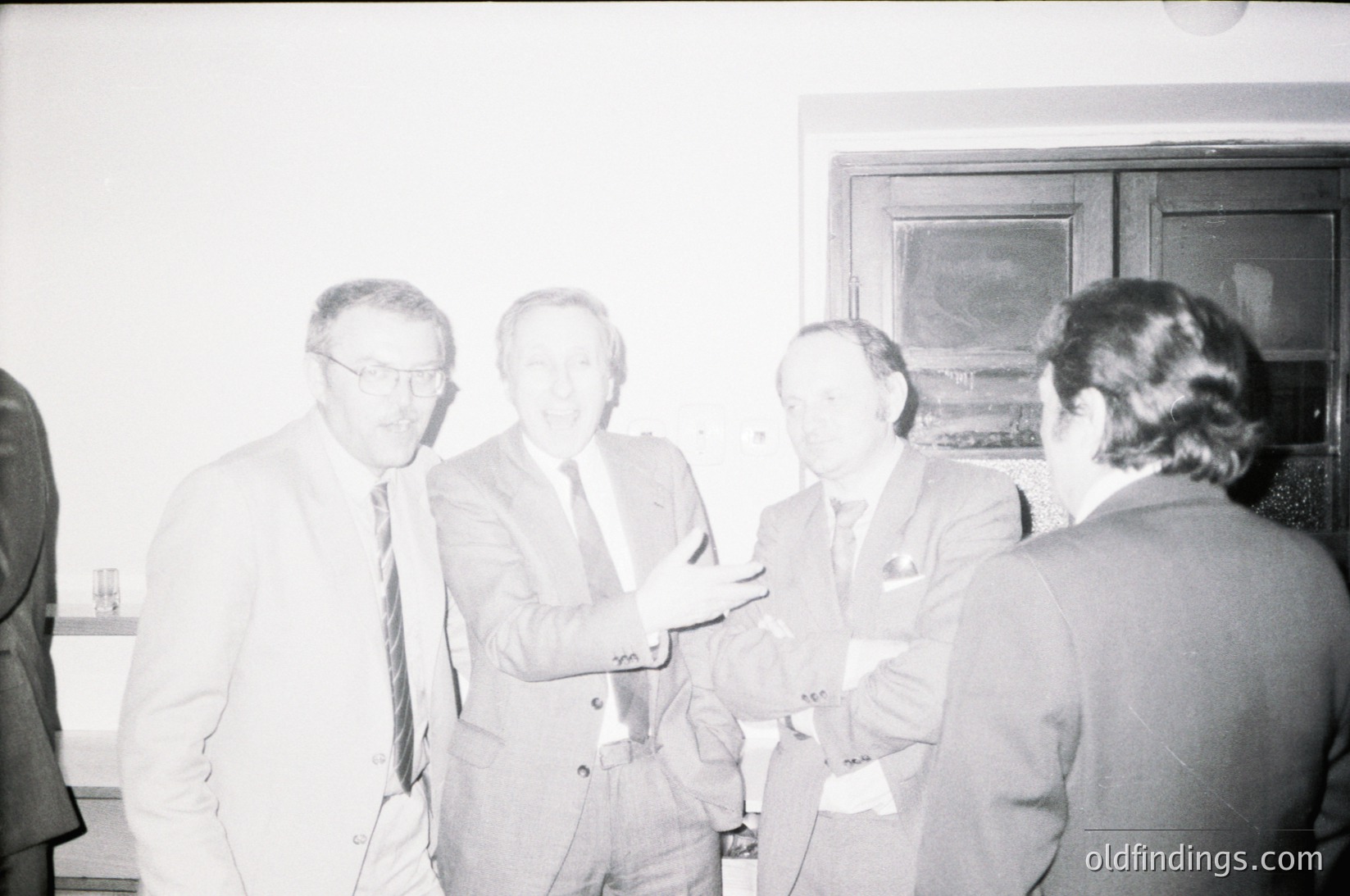 Four men in formal attire—dark suits, ties, and name badges—engage in conversation indoors, likely at a conference or professional event. Mid-20th century (1960s–1980s) setting with muted lighting and minimalist decor.