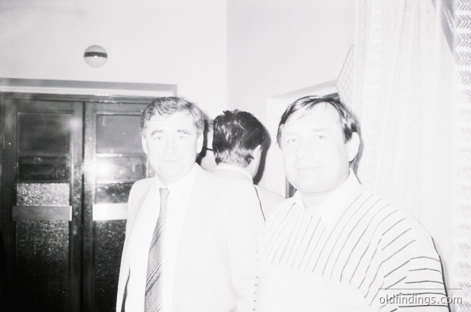 Black-and-white portrait of two men in formal attire, likely 1970s–1980s. The man on the left wears a suit with a patterned tie, while the man on the right sports a striped dress shirt and a cap. Background shows a blurred indoor setting with a refrigerator and curtain.