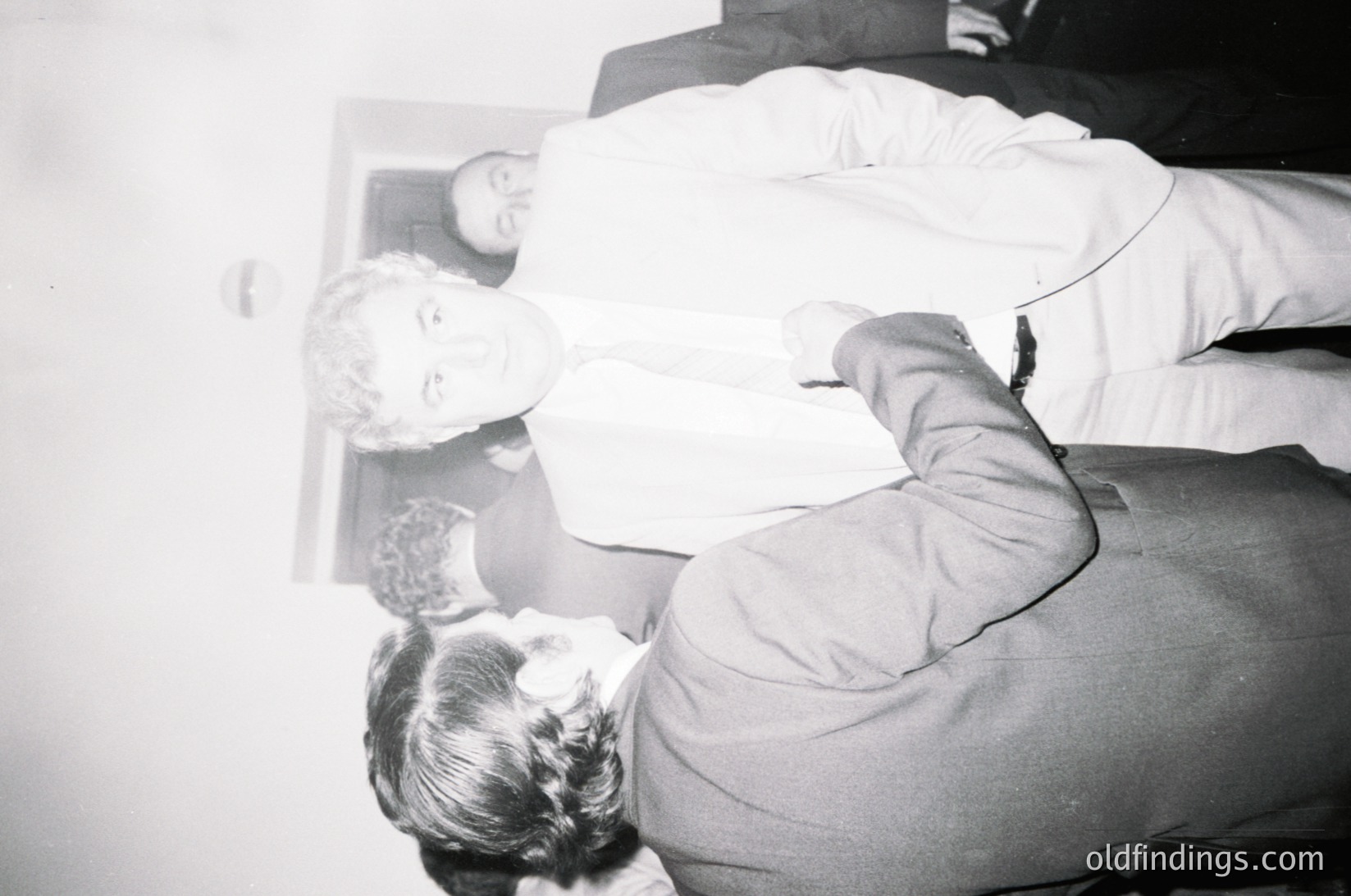 Vintage black-and-white photo of a seated man in formal attire, adjusting a woman’s hair in a mirror. Reflective surface reveals another man in a suit, suggesting an indoor event or portrait session. Mid-20th century styling () with classic grooming tools visible. Potential historical or fashion research value.