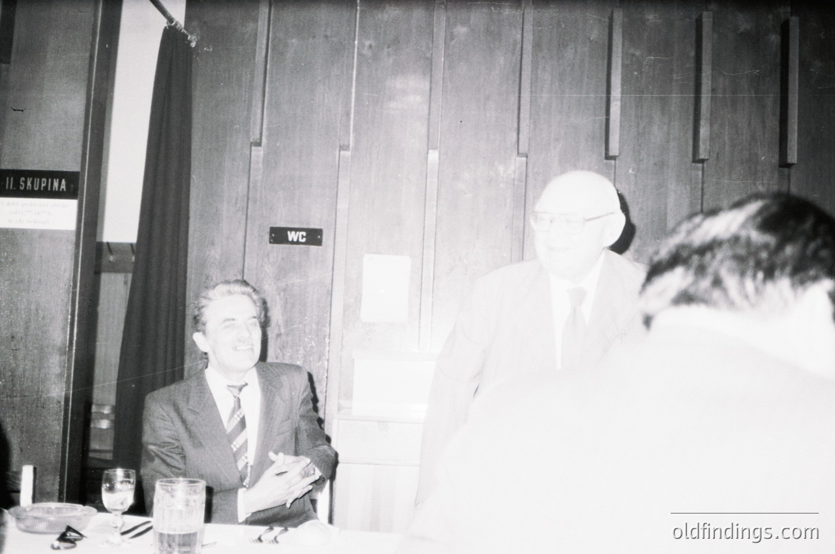 Black-and-white photo of three men in a formal indoor setting, likely a 1970s–1980s conference or meeting. Signage in Cyrillic ("WC" and "И СКУПИНА") suggests Eastern European location. Men wear suits, indicating professional or governmental context. Glasses, ashtrays, and a table with papers suggest a discussion or negotiation.