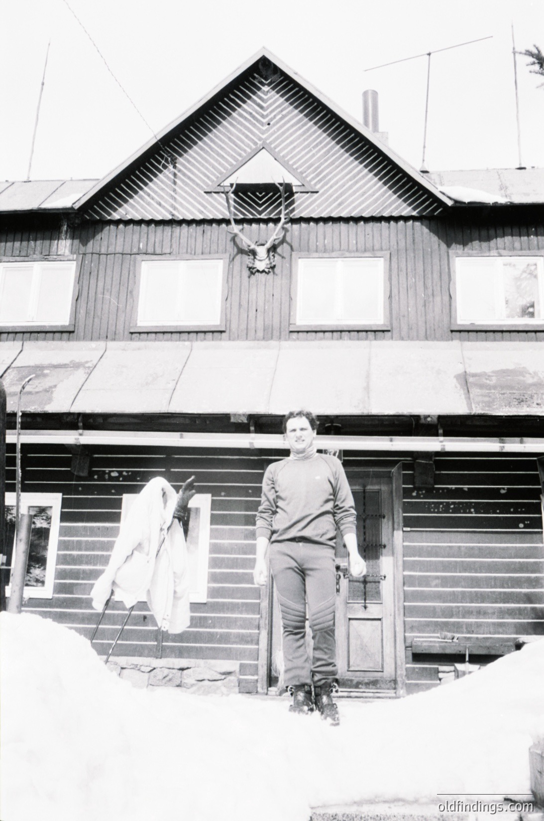 Classic black-and-white photo of a wooden log cabin in snowy terrain. Man in winter gear stands at entrance, holding a rifle. Snow-covered landscape and traditional alpine architecture suggest rural, remote setting. Likely 20th-century European mountain village.