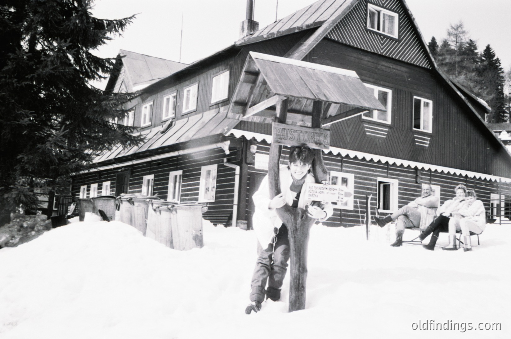 Two men in winter attire pose near a wooden alpine lodge with signage in Cyrillic. Snow blankets the ground and rooftops. One man holds a sign reading "Ски-център" (Ski Center).