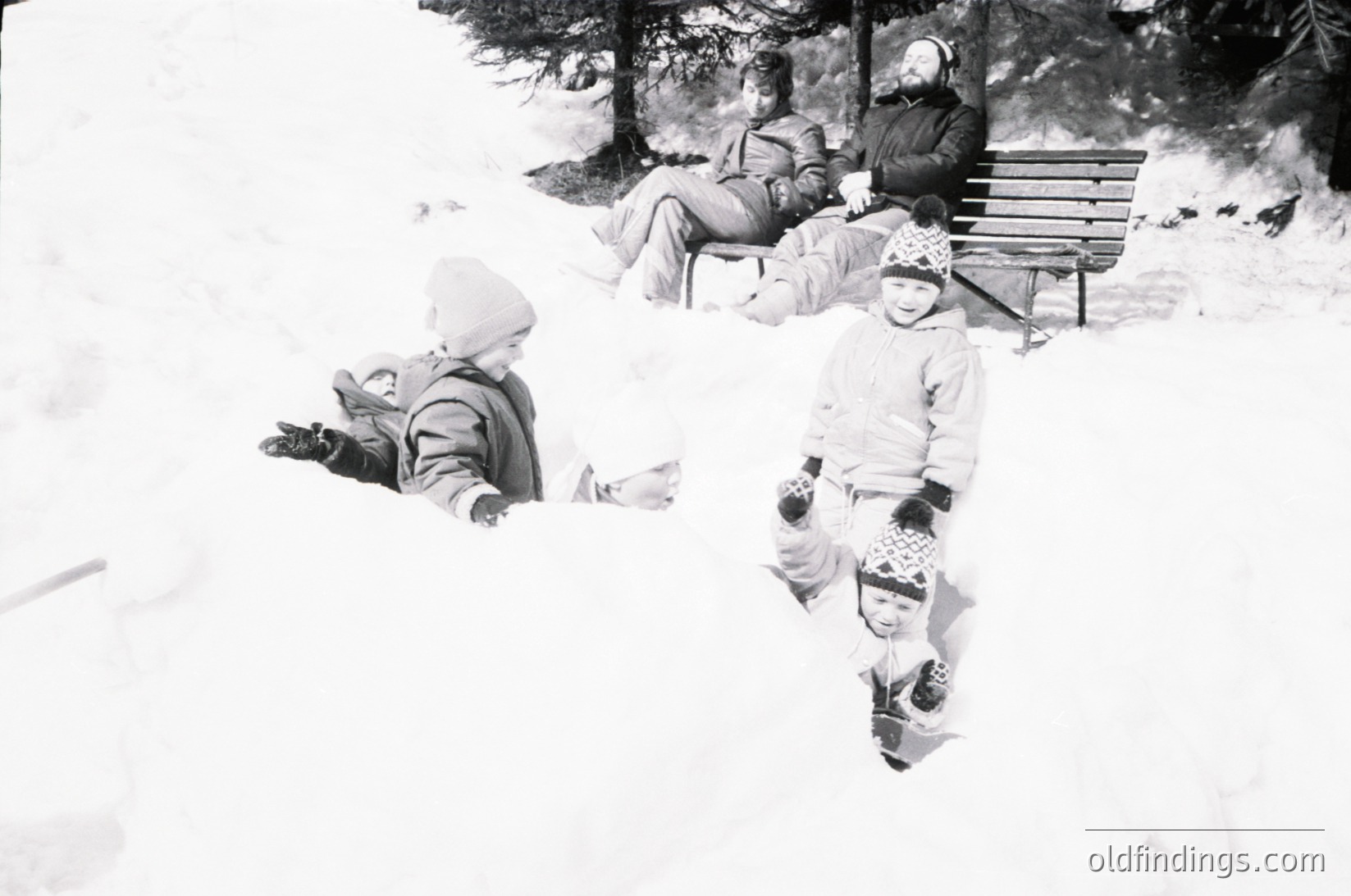 Mid-20th century black-and-white snapshot of winter family fun in alpine setting. Three children build snow structures while adults relax on a wooden bench under pine trees. Warm winter attire—knit hats, mittens, and scarves—suggests mid-century fashion. Likely taken in a European mountain region during the 1950s–1960s.