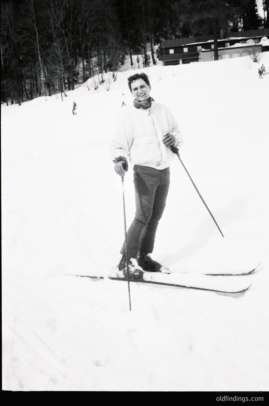 Black-and-white ski portrait of a person in mid-stance on groomed slopes, holding classic skis and poles. Forest and wooden structures visible in background, suggesting alpine resort. Clothing suggests 1960s–1970s skiwear.