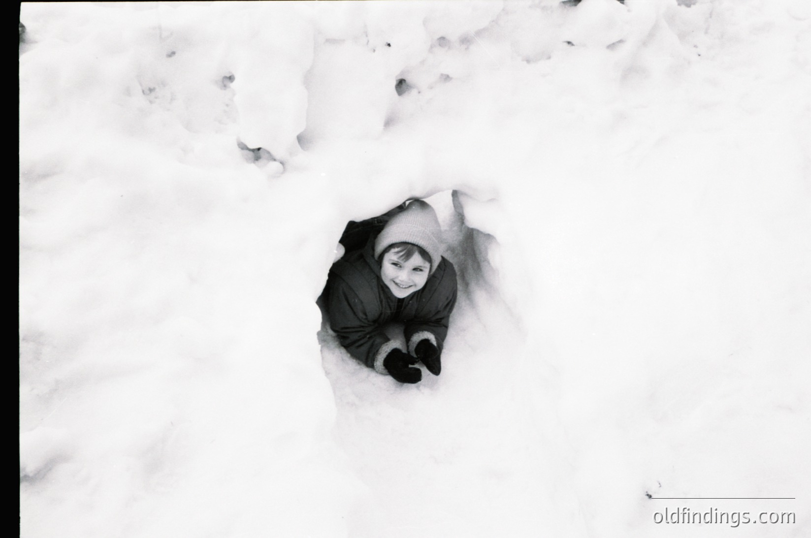 Black-and-white shot of a child buried up to their chest in snow, wearing a dark coat and hat. Playful winter scene evokes mid-20th century family life.