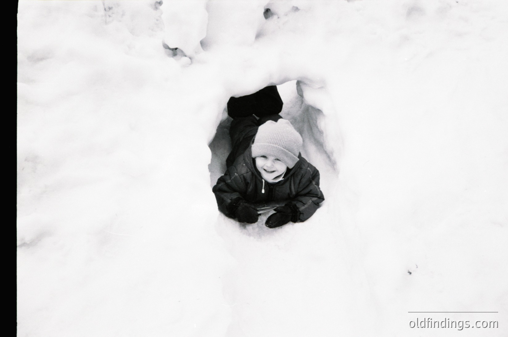 Child buried in snowdrift, smiling up from a dug-out hole. Black-and-white winter scene evoking mid-20th century play. Warm, candid expression contrasts with cold surroundings. Ideal for nostalgic or seasonal stock imagery.