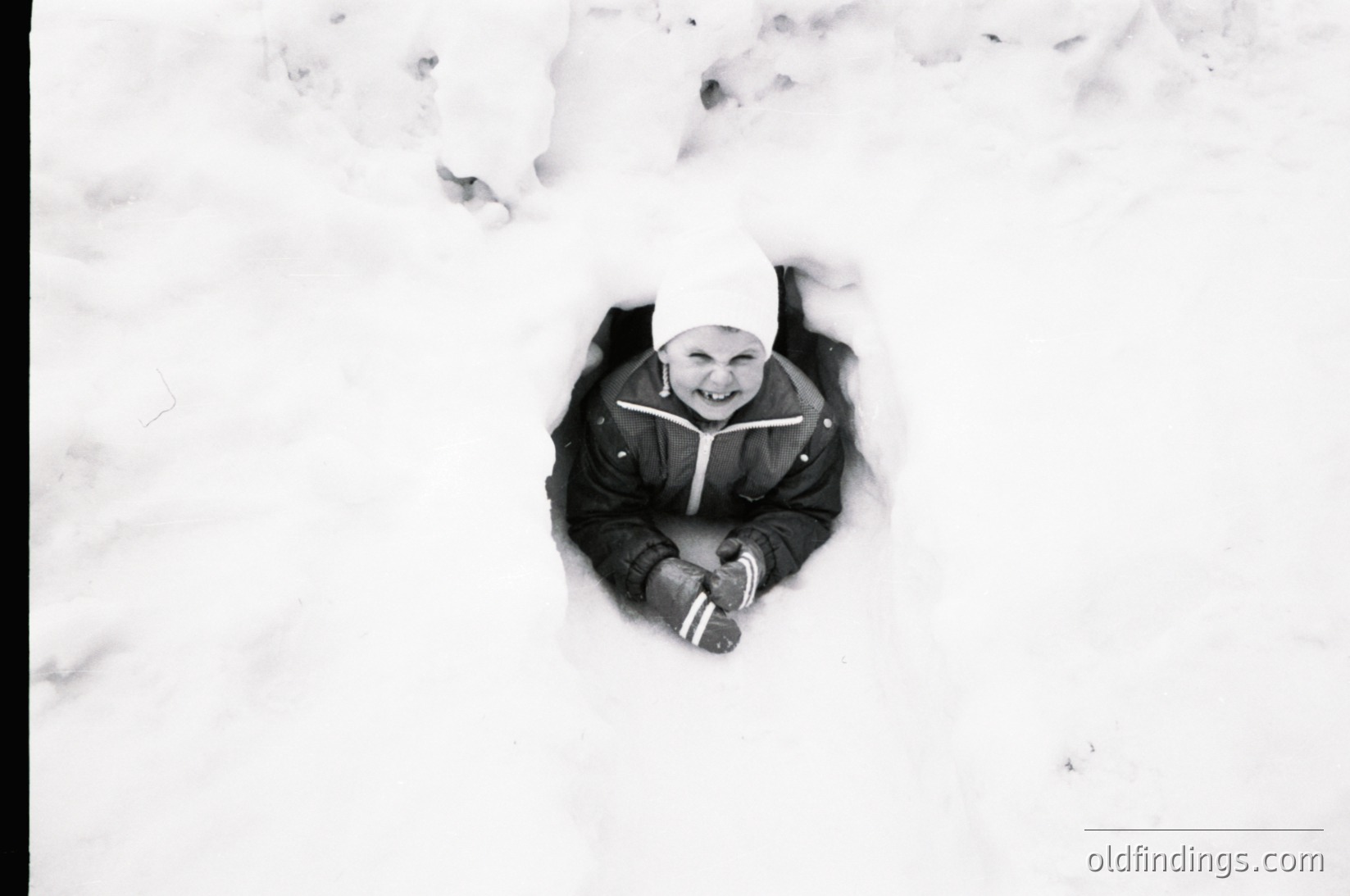 Child buried in snow, smiling up from a shallow hole, wearing a knit hat and winter coat. Black-and-white winter scene evoking mid-20th century play.