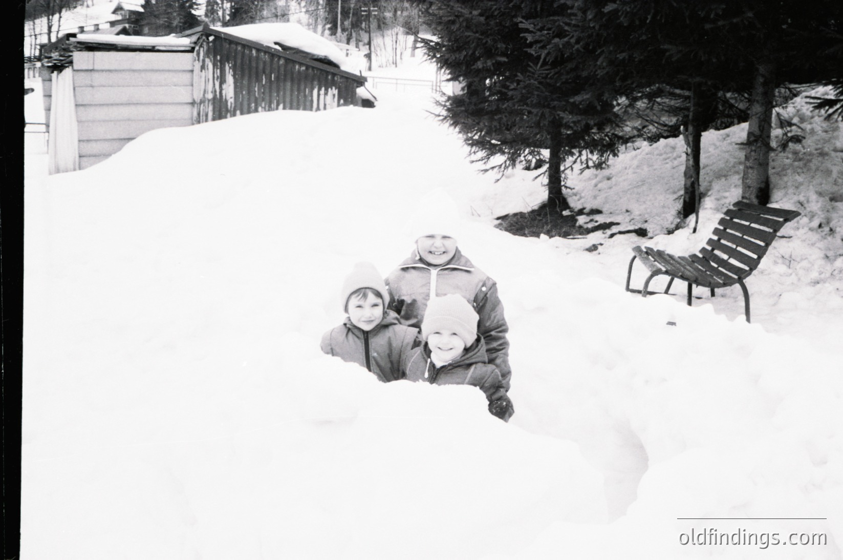 Three children pose playfully in deep snow, bundled in winter coats. Snow-covered alpine lodge and benches in background suggest a ski resort or mountain retreat. Black-and-white grain and lighting indicate mid-20th century (1950s–1970s) photography.