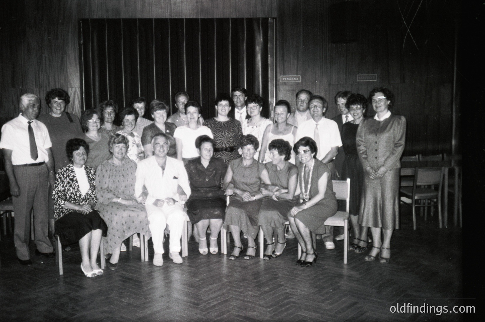 Group portrait of 20+ individuals in a dimly lit indoor setting, likely a community hall or theater. Seated on wooden chairs and benches, they wear 1960s–1970s fashion—blouses, cardigans, and patterned dresses. A sign in the background reads "САЛОН" (Salon). Formal, posed group shot with wooden flooring and vertical wooden panels.