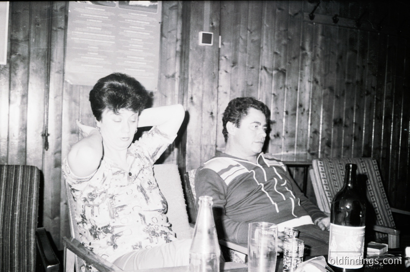 Two individuals seated in a rustic wooden interior, likely mid-20th century. Woman in floral dress leans back, man in striped shirt holds a wine bottle. Wooden plank walls and simple furnishings suggest a casual, informal setting. Possible domestic scene.