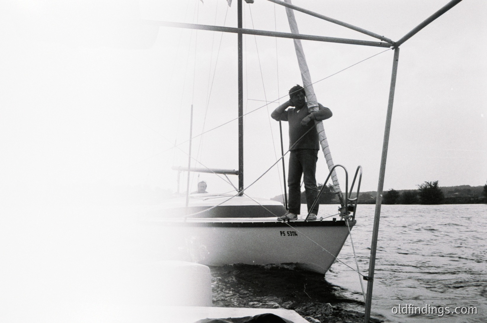 Black-and-white shot of a sailor adjusting a sail on a classic sailboat, marked "PS STIR." Mid-20th century maritime attire and vessel design suggest 1960s-1970s era. Overcast skies and calm waters imply coastal or inland lake setting.