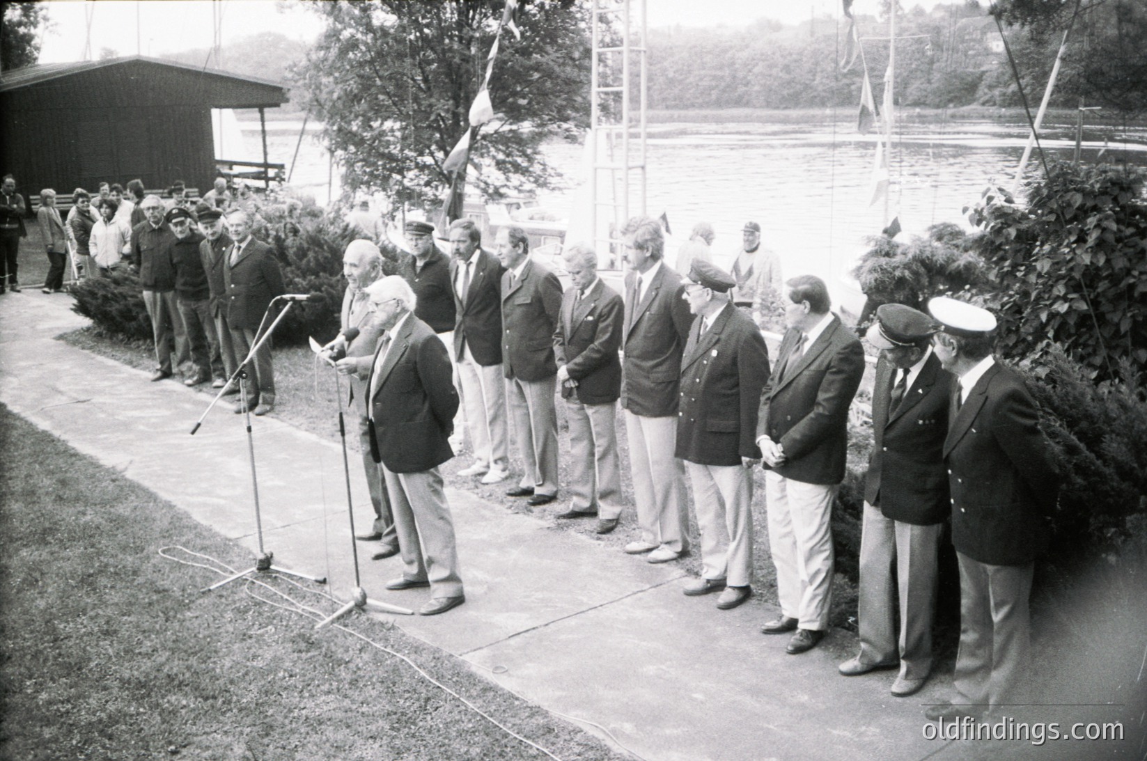 Mid-20th century formal ceremony with men in suits and caps standing in line for a speech or event. Microphone and trees in background suggest an outdoor venue, likely institutional or governmental.