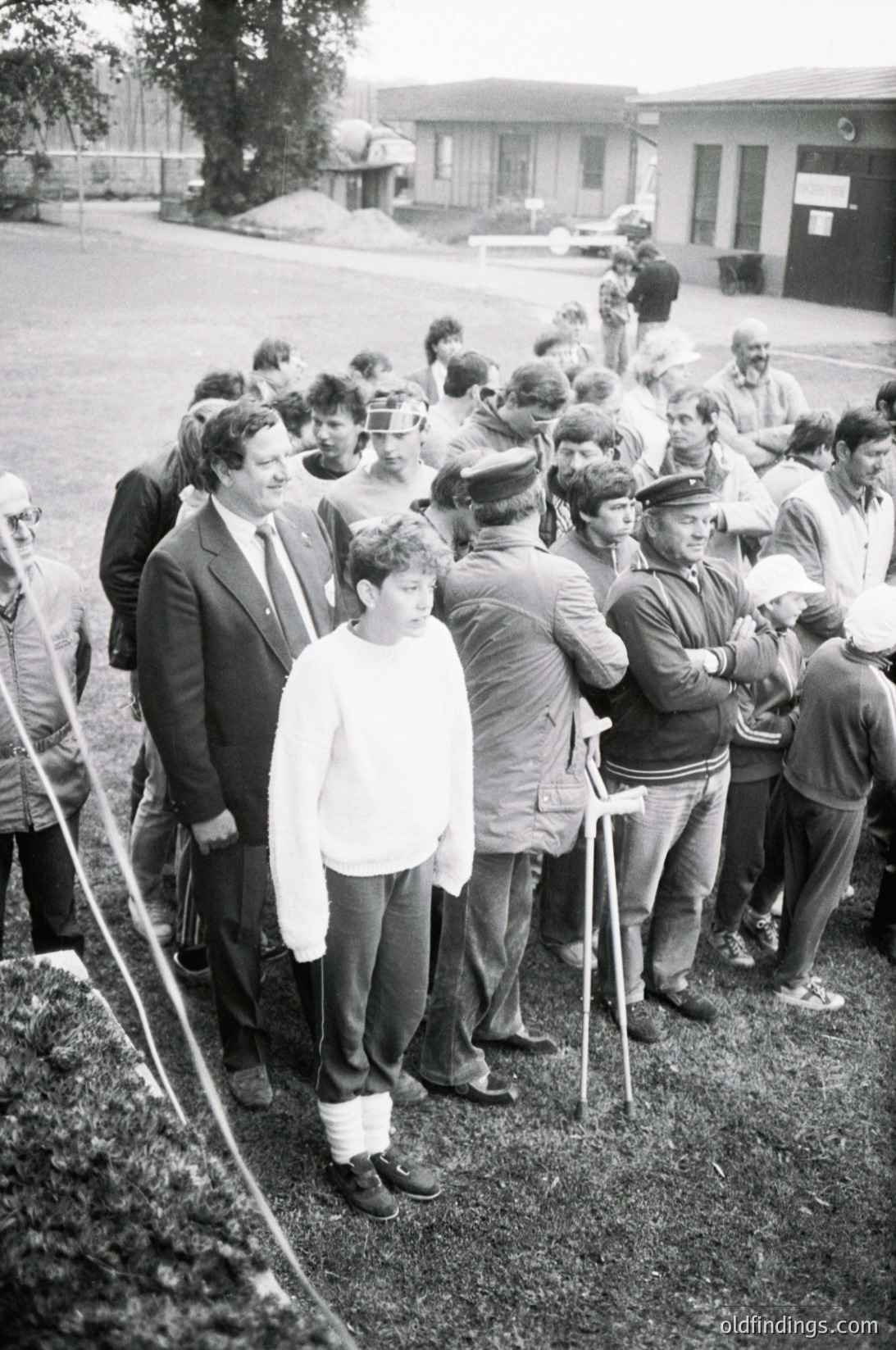 Black-and-white photo of a public gathering in an outdoor setting, likely a 1970s-1980s Eastern Bloc country. A diverse crowd, including men in suits and military-style caps, surrounds a central figure in a white shirt and dark pants. The scene suggests a formal event or demonstration, possibly political or civic. Concrete buildings and a paved area in the background.