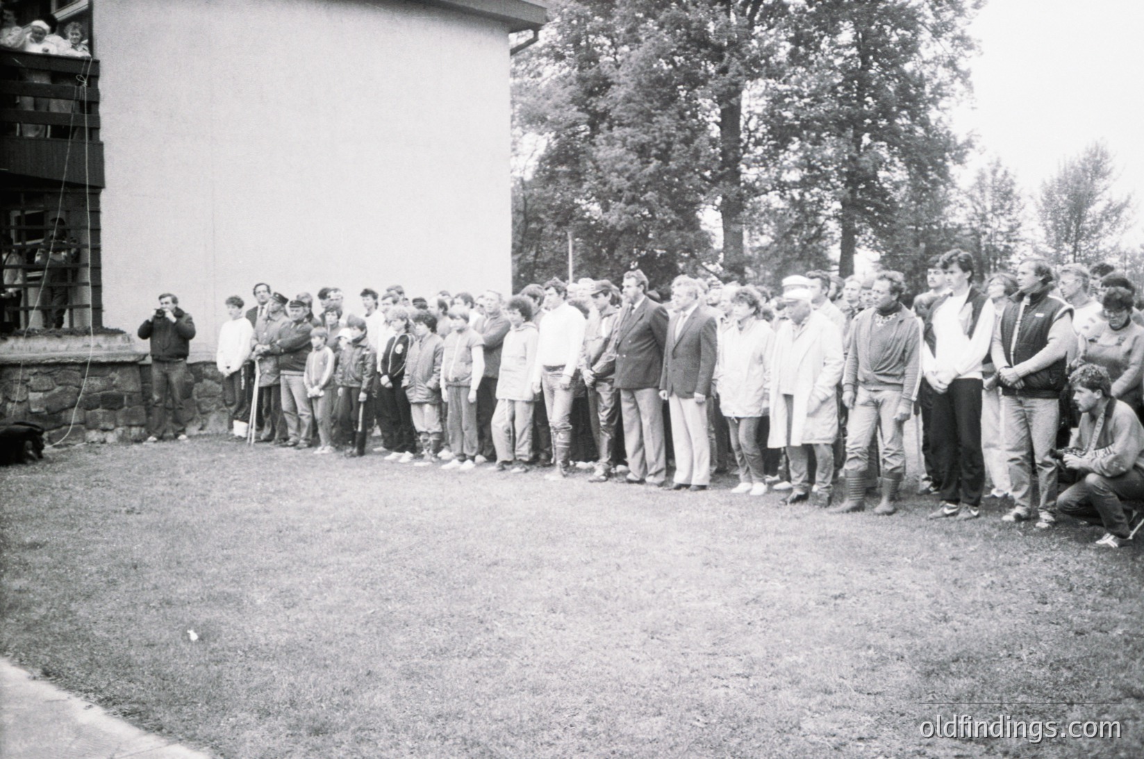 A black-and-white group photo of ~30 individuals standing in a single-file line outdoors, likely mid-20th century. Casual attire suggests a communal or work-related gathering, with some wearing caps and jackets. A building with scaffolding in the background indicates construction or renovation. Grass and trees frame the scene, suggesting a rural or suburban setting.