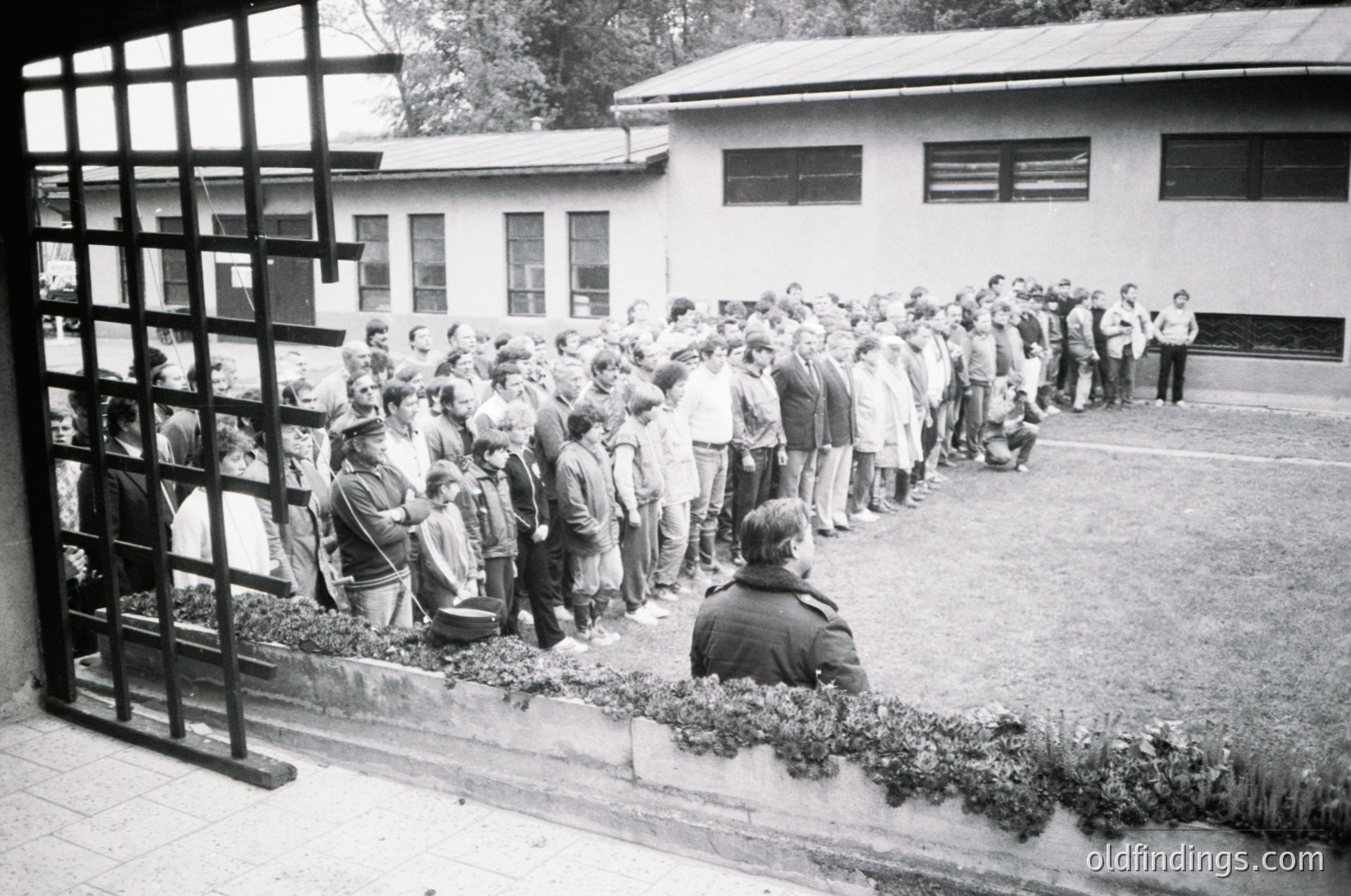 Long queue of people lined up outdoors, separated by a metal fence, near a mid-century institutional building. Uniform attire suggests a formal or organized event, likely mid-20th century.