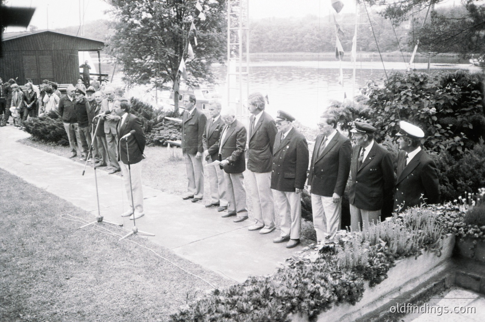 Black-and-white formal gathering of uniformed men in outdoor ceremony, likely mid-20th century. Men in suits, military caps, and peaked hats stand at attention near microphones and greenery. Background shows a crowd and a body of water, suggesting an official event or speech.