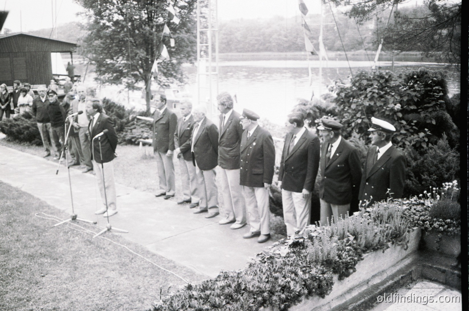 Black-and-white photo of formal outdoor gathering, likely mid-20th century. Group of men in suits, caps, and naval uniforms line up for a ceremony or inspection near a waterfront. Microphones and press cameras indicate media presence. Lush greenery and a distant bridge frame the scene.