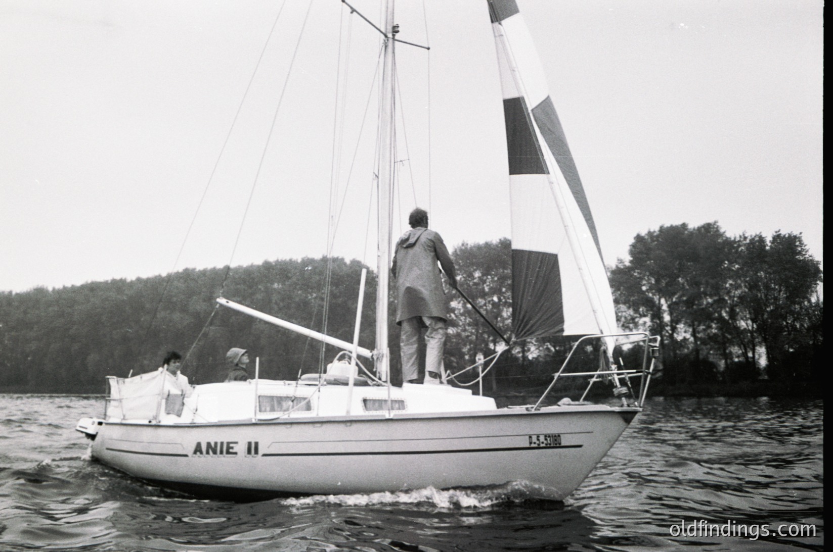 Classic 1970s sailboat "Anie II" cruising on calm waters, with two individuals aboard. The vessel features a white hull, tall mast, and striped sail. Lush forested shoreline in background suggests a serene lakeside or coastal setting.