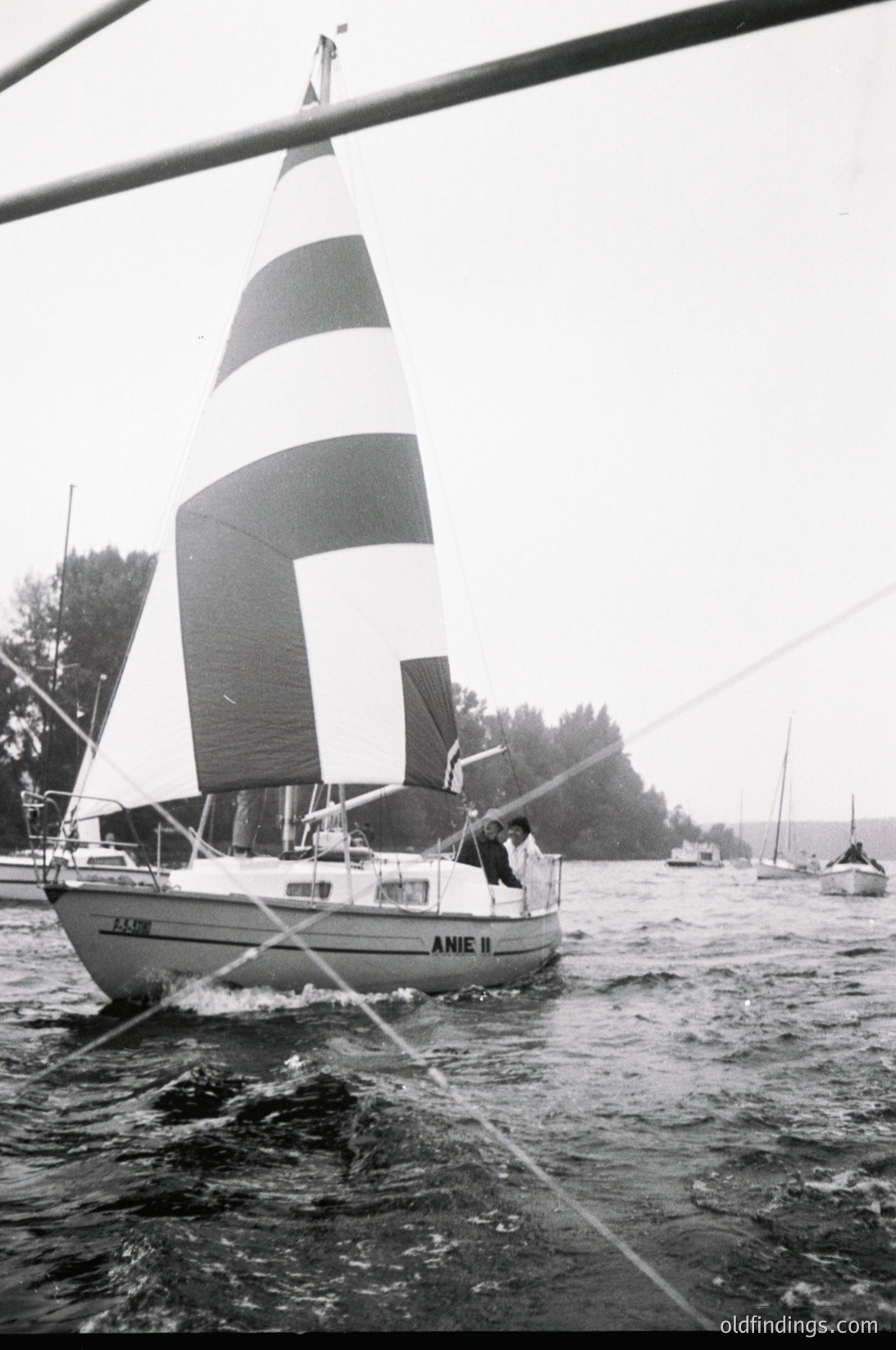 Classic 1970s sailboat "ANIE II" cutting through choppy waters, framed by a dock pole. Mid-century design with striped mainsail and white hull. Coastal marina or lake setting with other boats in background.
