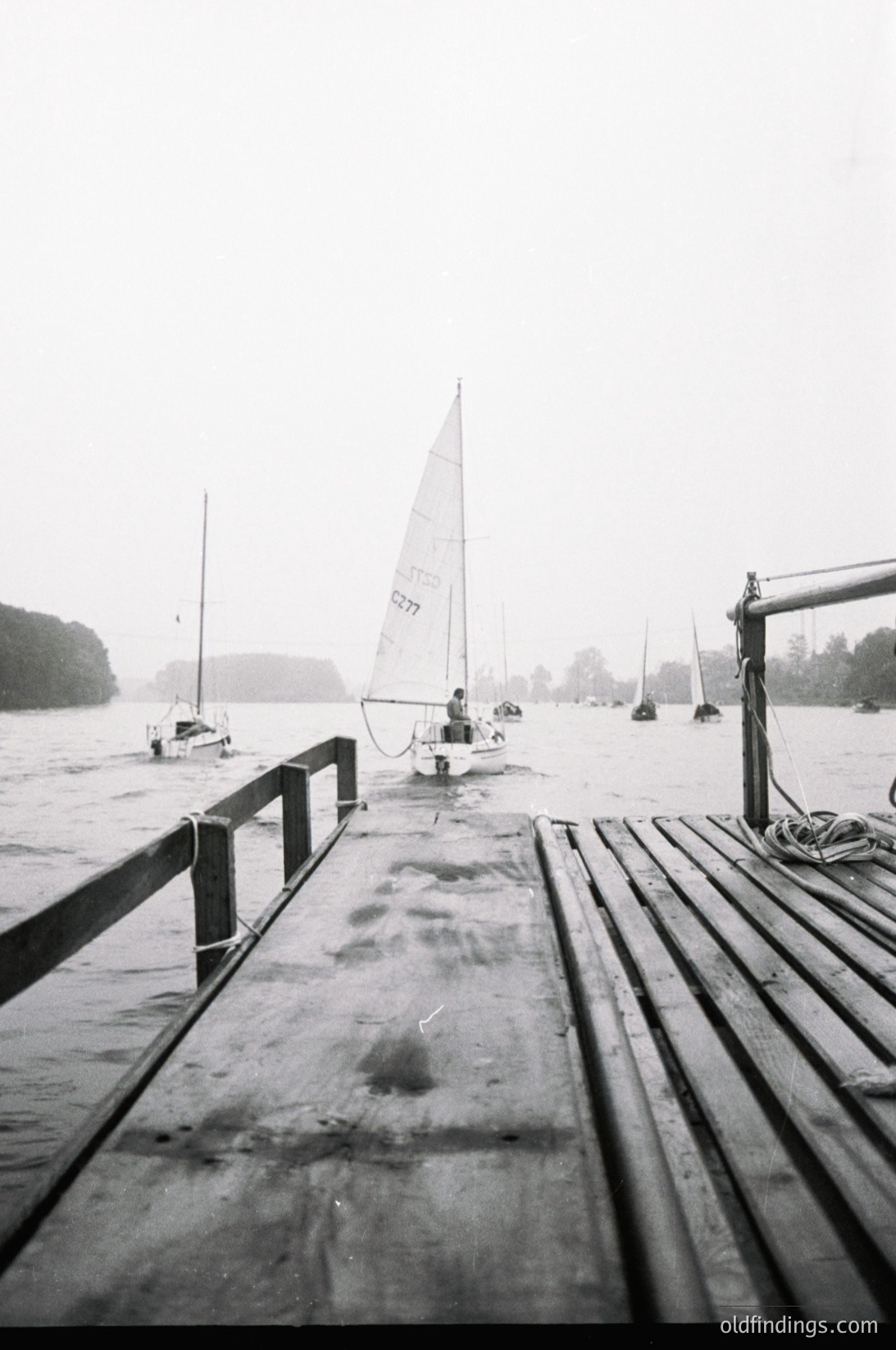 Black-and-white shot of a wooden dock extending into a misty lake, with a single-hander sailboat (number 8277) moored nearby. Overcast skies and distant trees frame the serene, early-morning scene. Mid-20th century maritime vibe.