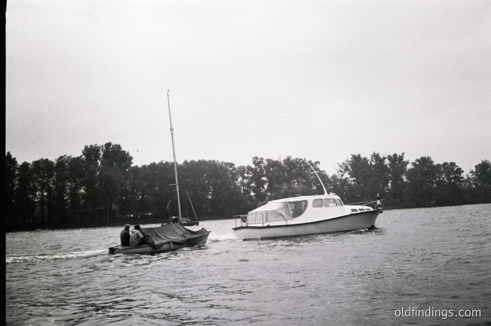 Vintage black-and-white photo of two small boats on calm waters: a motorized cabin cruiser and a simple sailboat with a covered deck. Dense tree line in background suggests a lake or river setting. Mid-20th century design reflects mid-century maritime aesthetics.