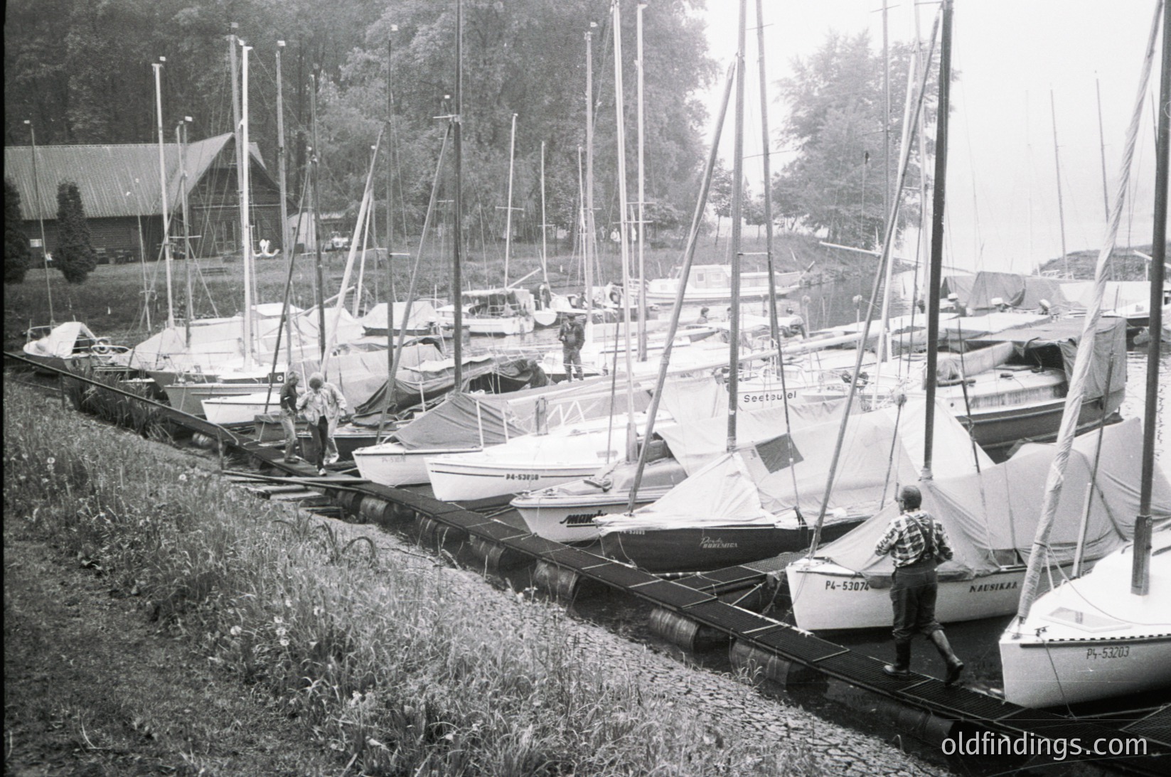 Classic black-and-white marina scene featuring a row of small sailboats and motorboats docked on wooden supports. A man in a striped sweater stands near the foreground, while others inspect or work on vessels. Wooden pier and lush greenery frame the scene, suggesting a lakeside or coastal setting. Likely mid-20th century (1950s–1970s).