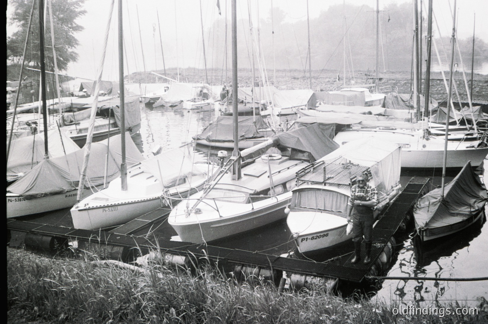 Docked vintage sailboats in a marina, likely mid-20th century. Classic fiberglass hulls with simple rigging and covered cockpits, some bearing registration numbers. Overcast skies and calm water suggest a coastal or lakeside setting. --- *Note: Exact location/time indeterminate but stylistic cues align with mid-20th-century recreational boating.*