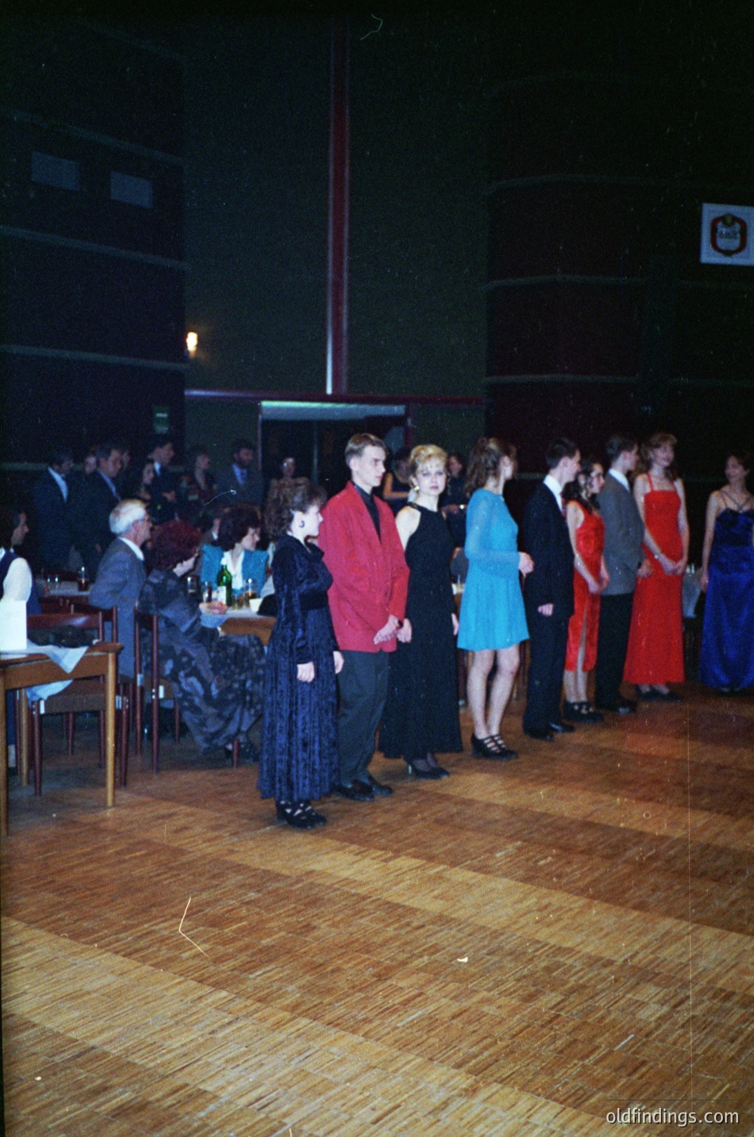 Vintage indoor dance event featuring 1970s-era attire: men in suits, women in floor-length dresses. Wooden dance floor with polished planks. Crowd seated at tables along walls, some holding drinks. Dim lighting with stage lighting visible. Likely a formal or semi-formal gathering.