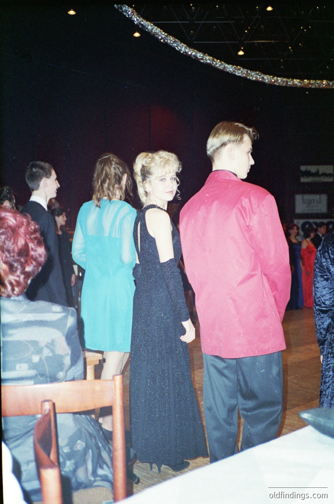Prom-style formal event with blurred crowd in background. Central figures in 1980s-era attire: a woman in a sleeveless black dress with a high slit, a man in a pink button-up shirt and jeans, and another in a teal dress. Indoor venue with stage lighting and hanging decorations. Likely a high school or youth formal.