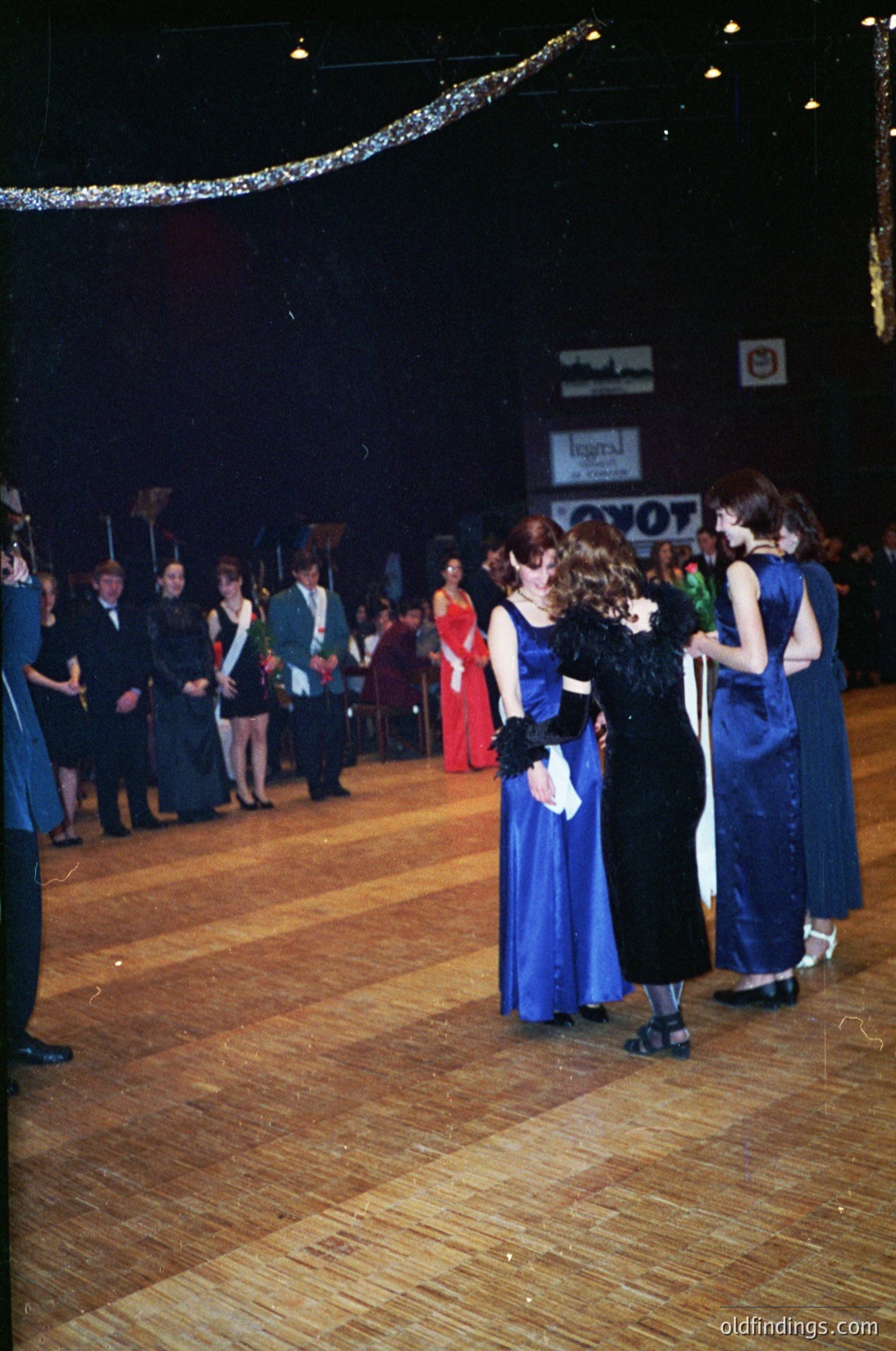 Vintage school prom scene with students in 1970s formal attire—mixed blue/green dresses, black suits, and bow ties. Wooden dance floor under a tinsel arch. Prom banner reads "Loyola" with "VOY" partial text. Crowded hall with dim lighting and a stage backdrop.