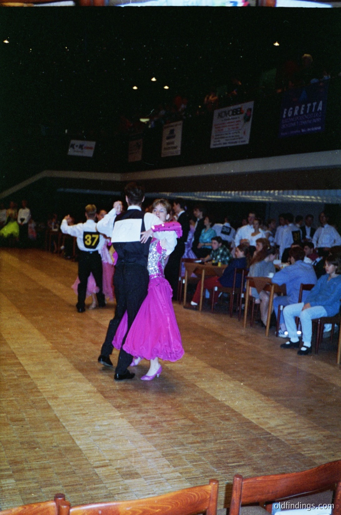 Couple ballroom dancing in a dimly lit hall, likely 1980s–1990s. Man in black suit, woman in pink ballgown. Spectators seated at wooden tables with numbered signs (, ). Advertisements for "Igretta" and "Jokebs" visible. Indoor social event setting.