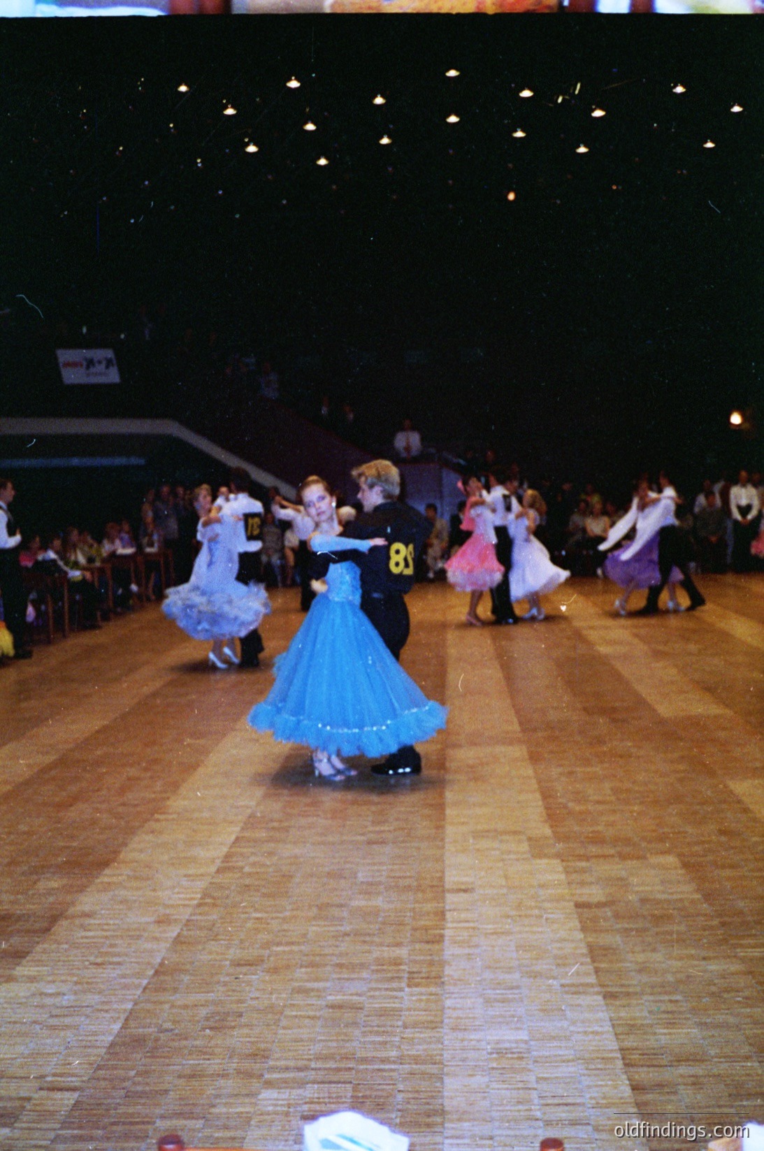 Group ballroom dance competition in a dimly lit hall, featuring children in formal attire. Boys wear suits with numbered bibs (, ), girls in voluminous skirts and blouses. Spectators seated in tiered bleachers. Likely 1980s–1990s Eastern European dance event.