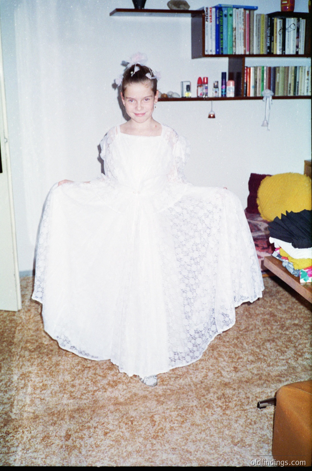 Vintage indoor portrait of a young girl in a voluminous lace wedding dress, posed on a patterned carpet. Bookshelves with colorful hardcover books and a plush toy in the background suggest a mid-20th century home setting. [Vintage 1950s-60s indoor portrait of girl in lace wedding dress, surrounded by bookshelves and plush toys ]