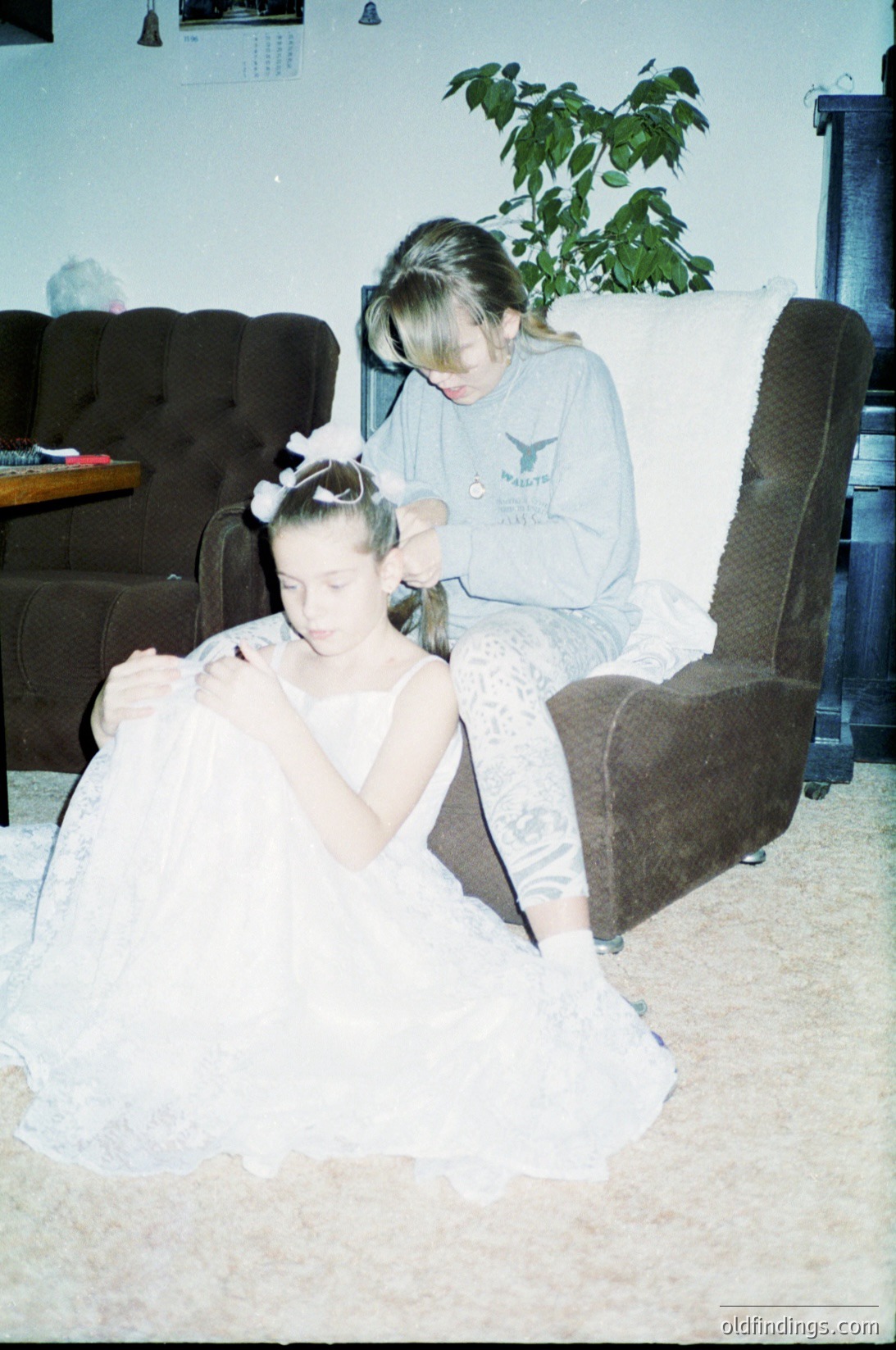 Vintage indoor portrait of two women in a living room setting, likely mid-1990s. One woman assists another in dressing a white, sleeveless gown, possibly for a formal event. The older woman wears a grey hoodie with a graphic print and a headband, while the younger woman sits in a brown armchair. A potted plant and a wooden coffee table with a framed photo are visible. Warm, nostalgic lighting enhances the candid moment.