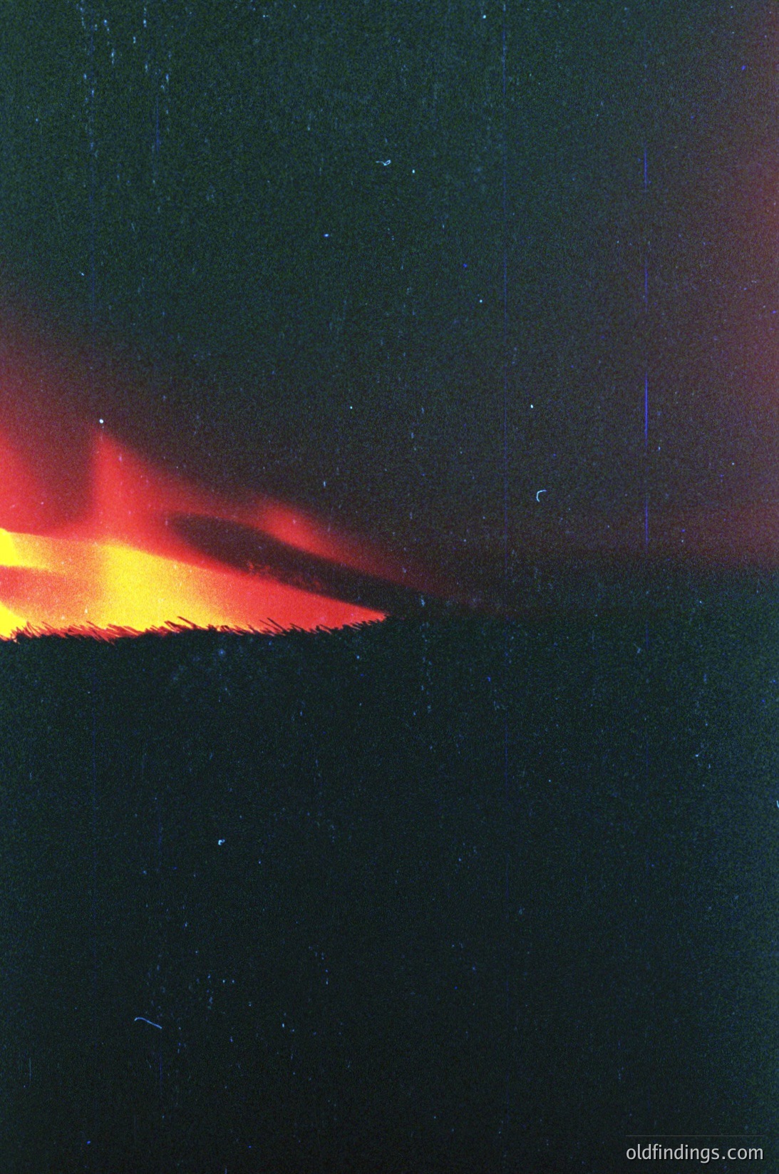Vivid red-orange flames erupting against a dark, textured background, likely a brick or concrete wall. The fire appears contained within a narrow, horizontal opening, suggesting a controlled burn or industrial setting. The grainy, muted tones suggest an older photograph, possibly from the mid-to-late 20th century. The composition highlights dramatic contrast and industrial aesthetics.