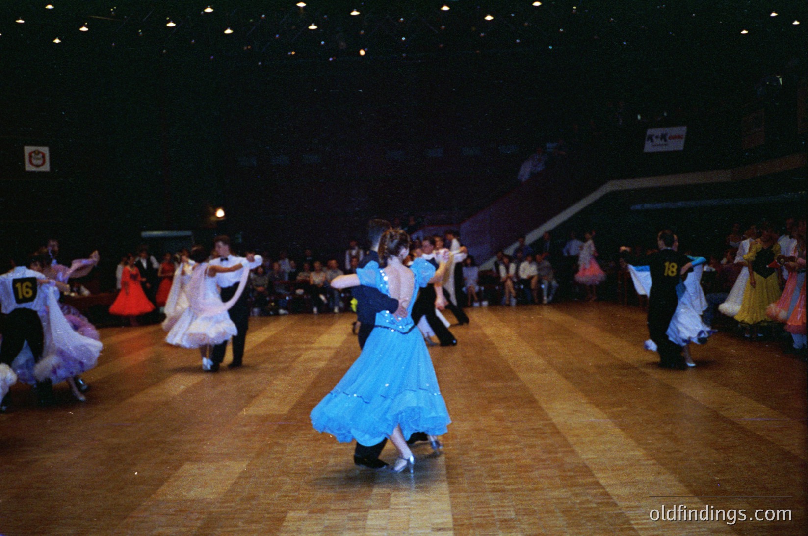 Indoor ballroom dance competition featuring couples in traditional folk attire. Men in dark suits with numbered bibs (, ) paired with women in vibrant, layered dresses (blue, white, red). Wooden dance floor under bright stage lighting. Crowd seated in tiered bleachers, suggesting a formal event. Likely Eastern European folk dance style, 1960s–1980s era.