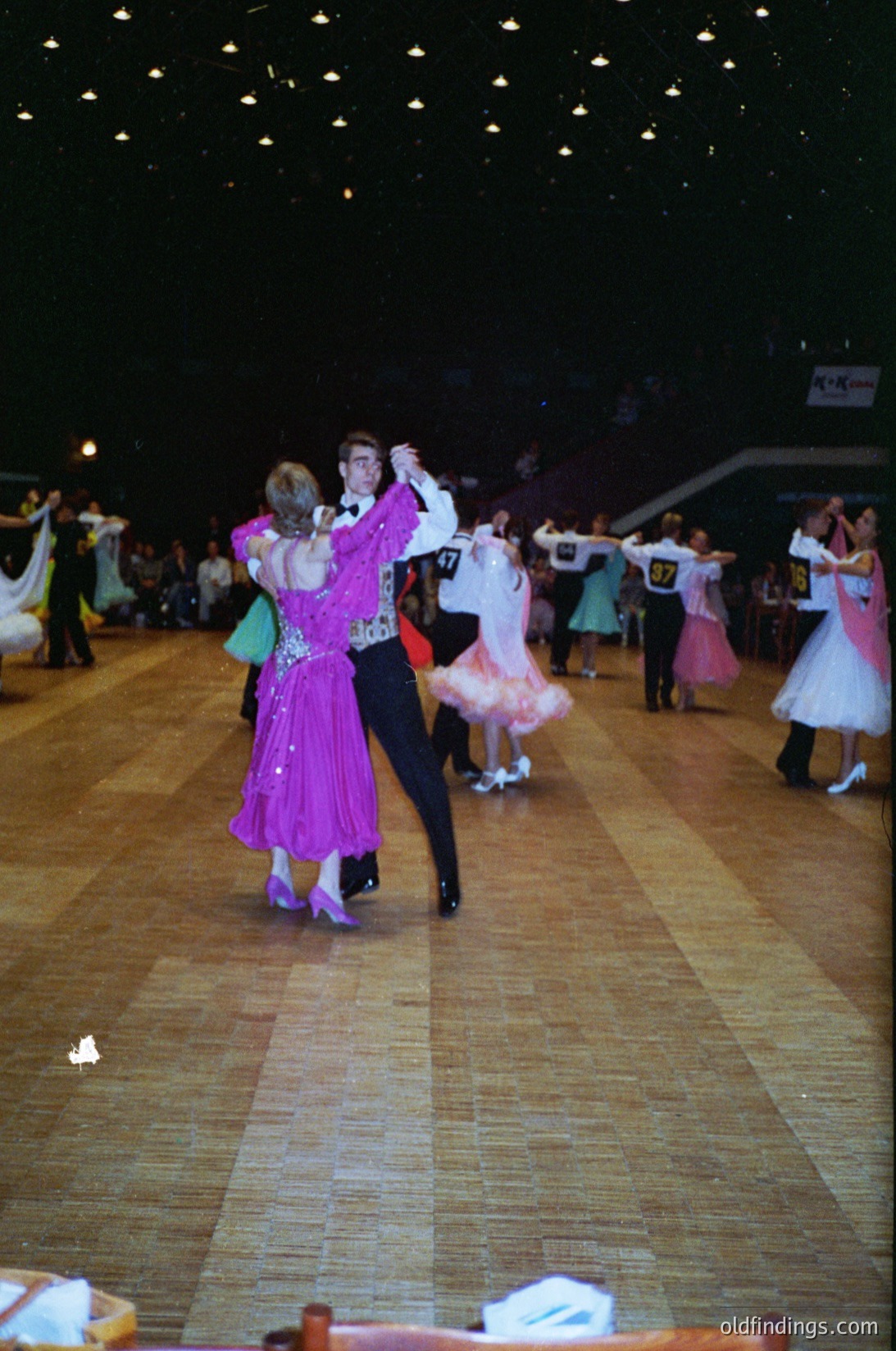 Indoor folk dance performance in a grand hall, likely 1980s–1990s Eastern Europe. Couples in traditional attire—women in flowing skirts with embroidered vests, men in tailored suits—dance under string lights. Wooden floor and tiered seating suggest a cultural festival or community event.