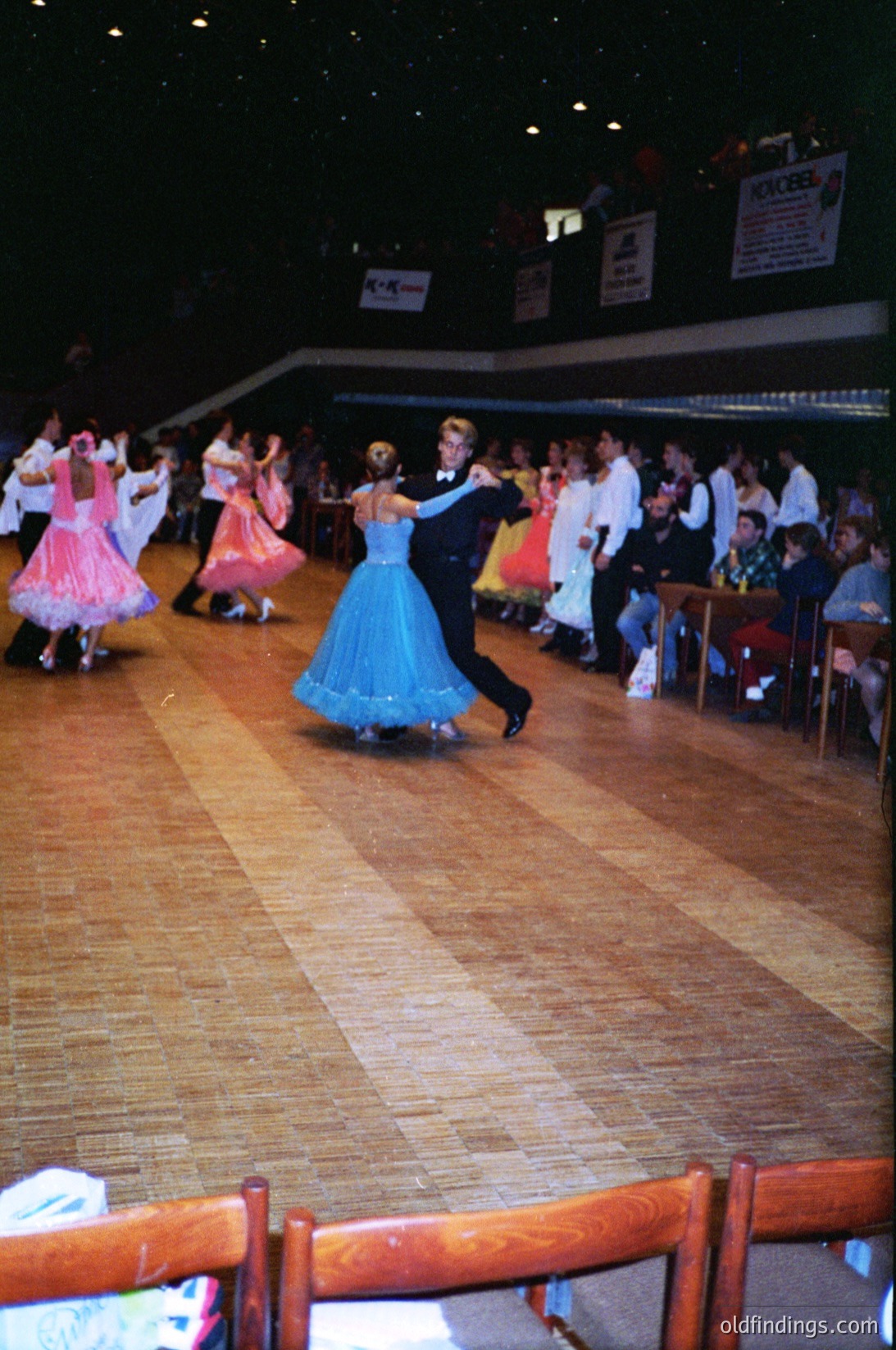 Ballroom dancers in 1970s-style attire perform on polished wooden floor. Couple in blue and black formalwear leads; spectators seated at wooden tables. Indoor venue with stage backdrop and signage.