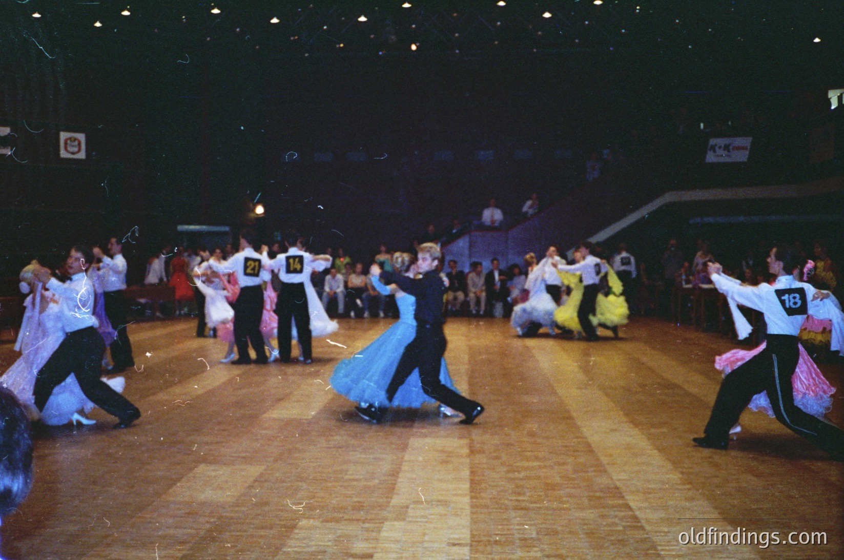 Ballroom dance competition featuring couples in 1970s-style attire—men in white jackets, black pants, and numbered bibs; women in flowing dresses (blue, yellow, pink). Indoor venue with wooden floor and spectators seated/standing.