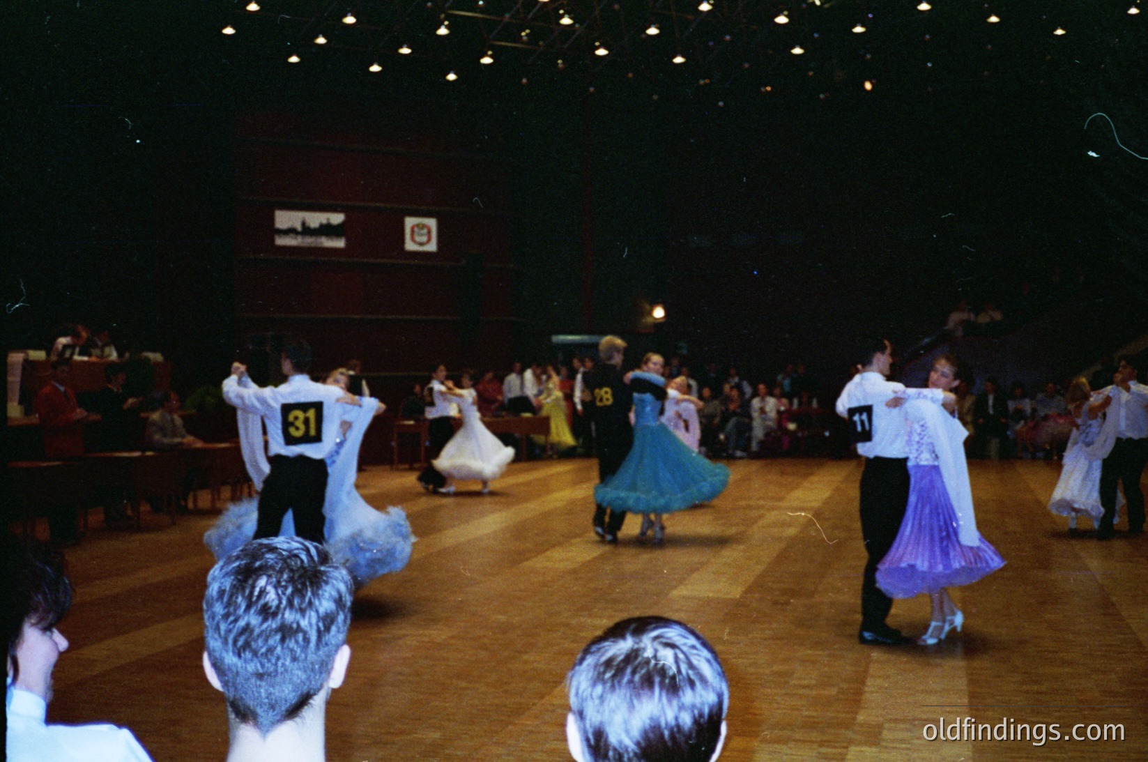 Indoor folk dance performance in a large hall, likely from the 1970s–1980s. Couples in traditional attire—men in white shirts with numbered bibs (, ) and dark pants, women in colorful skirts (blue, purple)—dance in a circular formation. Spectators seated along the perimeter observe. Warm lighting and polished wooden floor enhance the festive atmosphere.