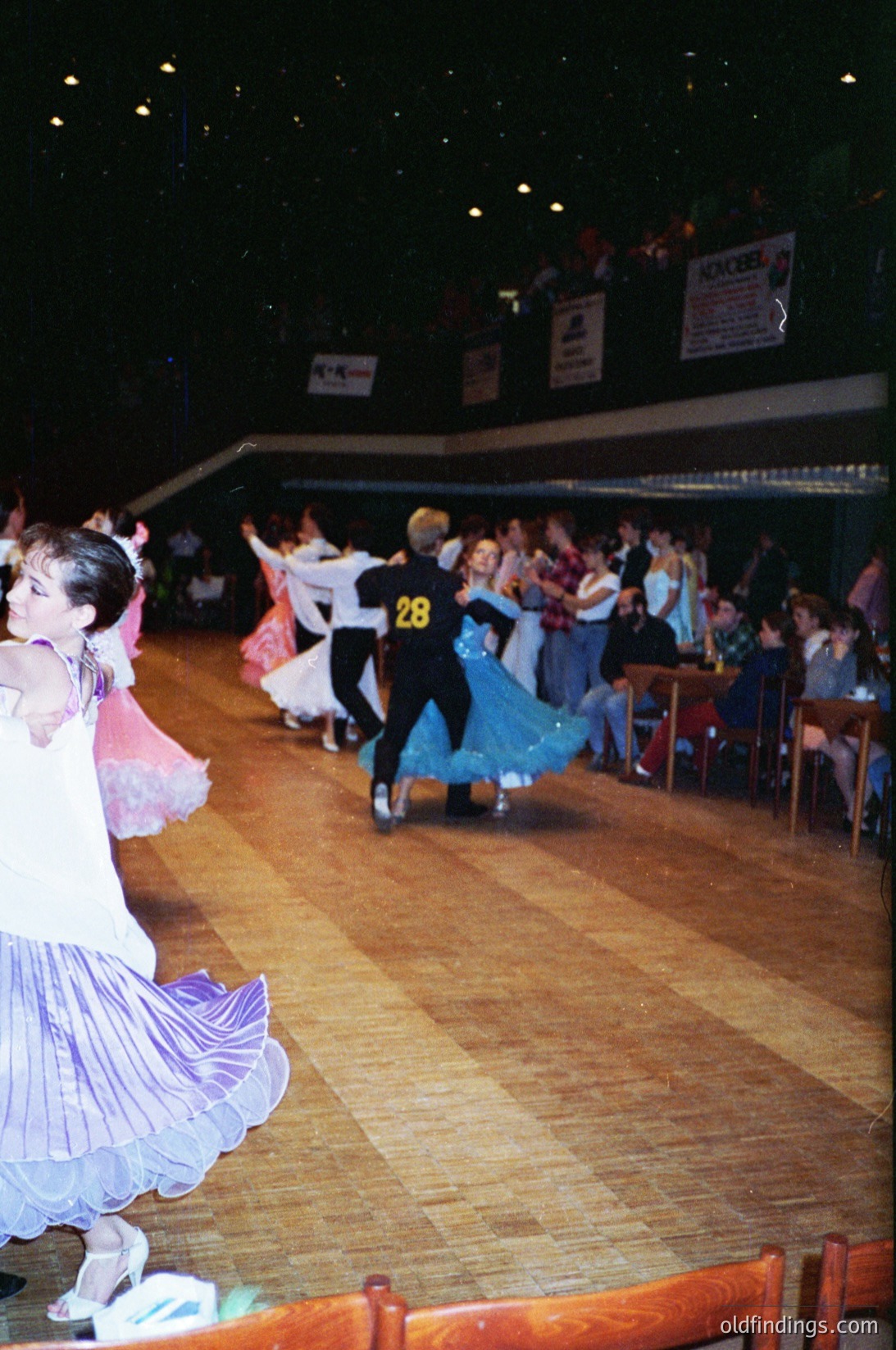 Ballroom dancers in 1970s-style attire perform indoors, likely at a competition or social event. Wooden floor, tiered seating, and banners suggest a formal venue. Competitors wear numbered vests ( visible) and elaborate skirts/leotards. Crowd watches from bleachers.