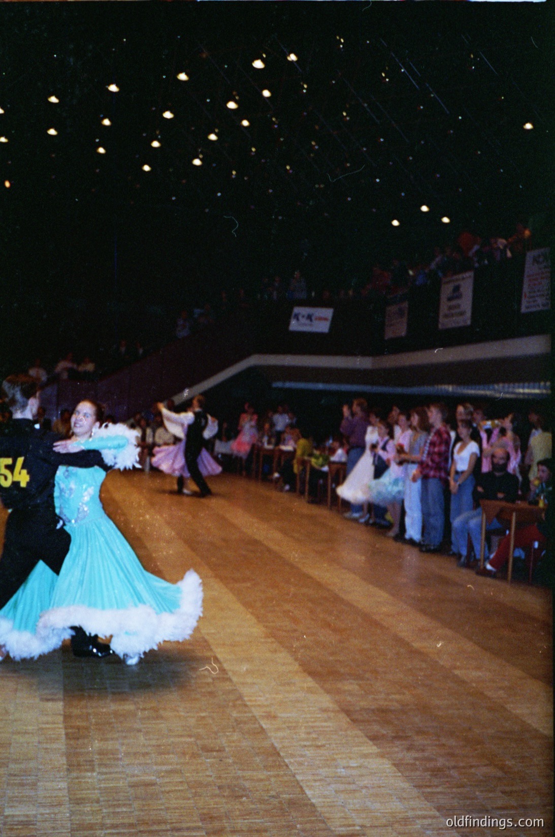 Ballroom dancers in 1970s-style attire perform under string lights in a spacious indoor venue. The woman wears a turquoise gown with white trim; the man sports a black suit with a numbered tag (). Crowd of spectators lines the perimeter, seated on bleachers. Decorated with banners and signs, likely a competition or social event.