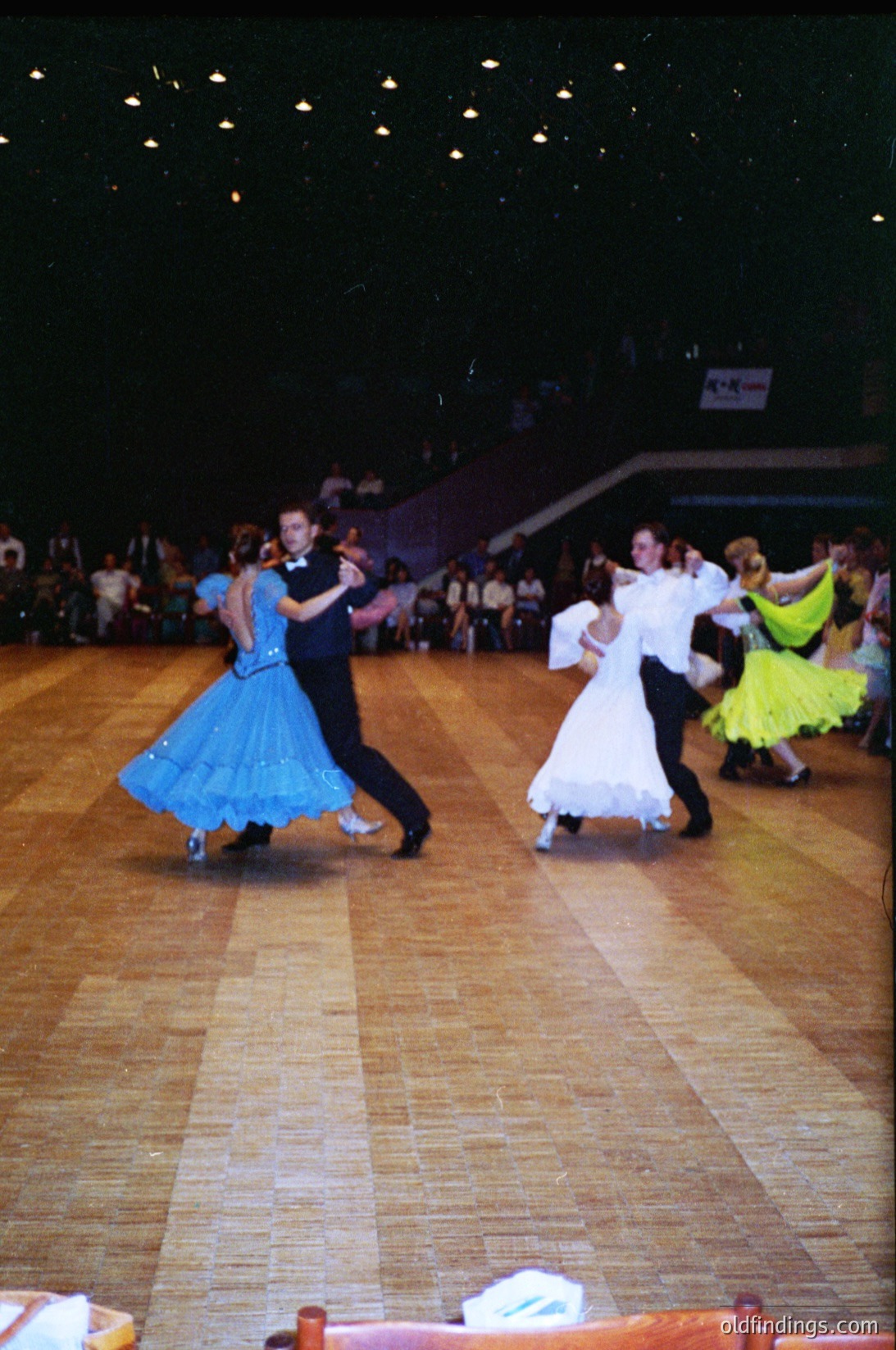 Ballroom dancers in vintage attire perform on polished wooden floors under stadium lighting. Men in dark suits paired with women in colorful, full-skirted dresses (, ). Crowd seated in tiered bleachers suggests an indoor venue, likely a cultural or recreational center. Authentic retro aesthetic ideal for historical research or nostalgic design references.