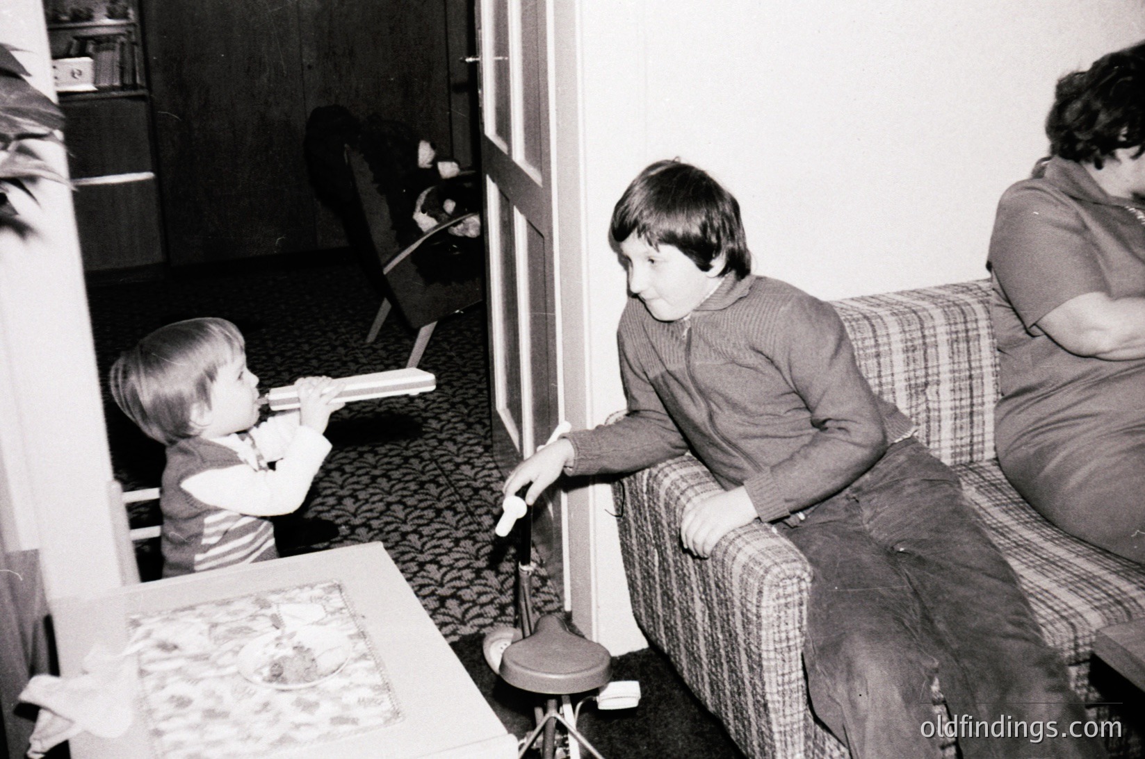 Vintage black-and-white photo of two children playing with a toy train set on a small table, supervised by an adult. Mid-20th century domestic interior with patterned carpet and wooden furniture.