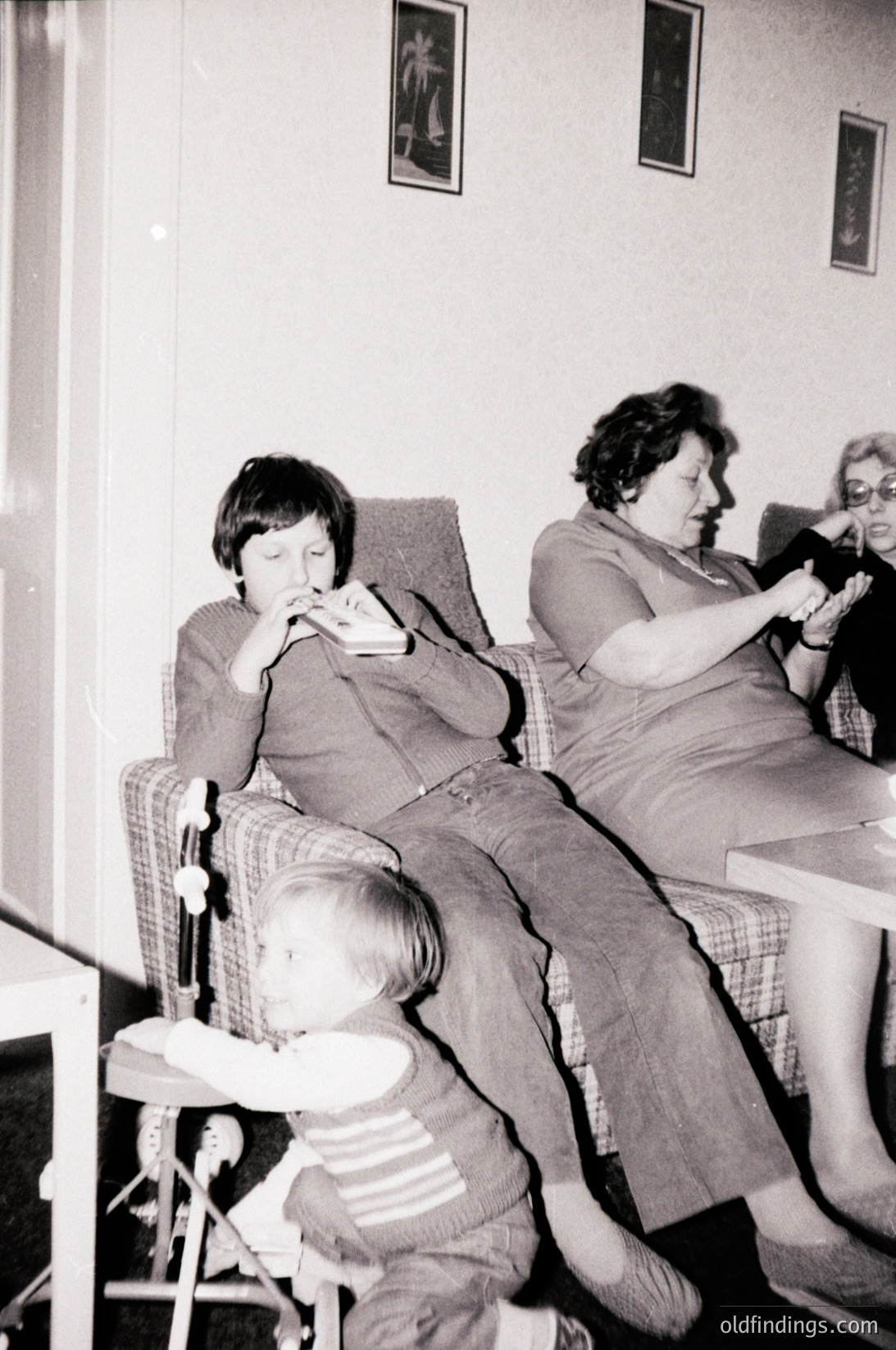 Vintage black-and-white indoor scene featuring three generations: a young boy eating, an adult woman knitting, and a toddler seated on a highchair. Mid-century furniture and framed tropical prints on walls suggest a 1960s–1970s domestic setting. Warm, candid family moment.