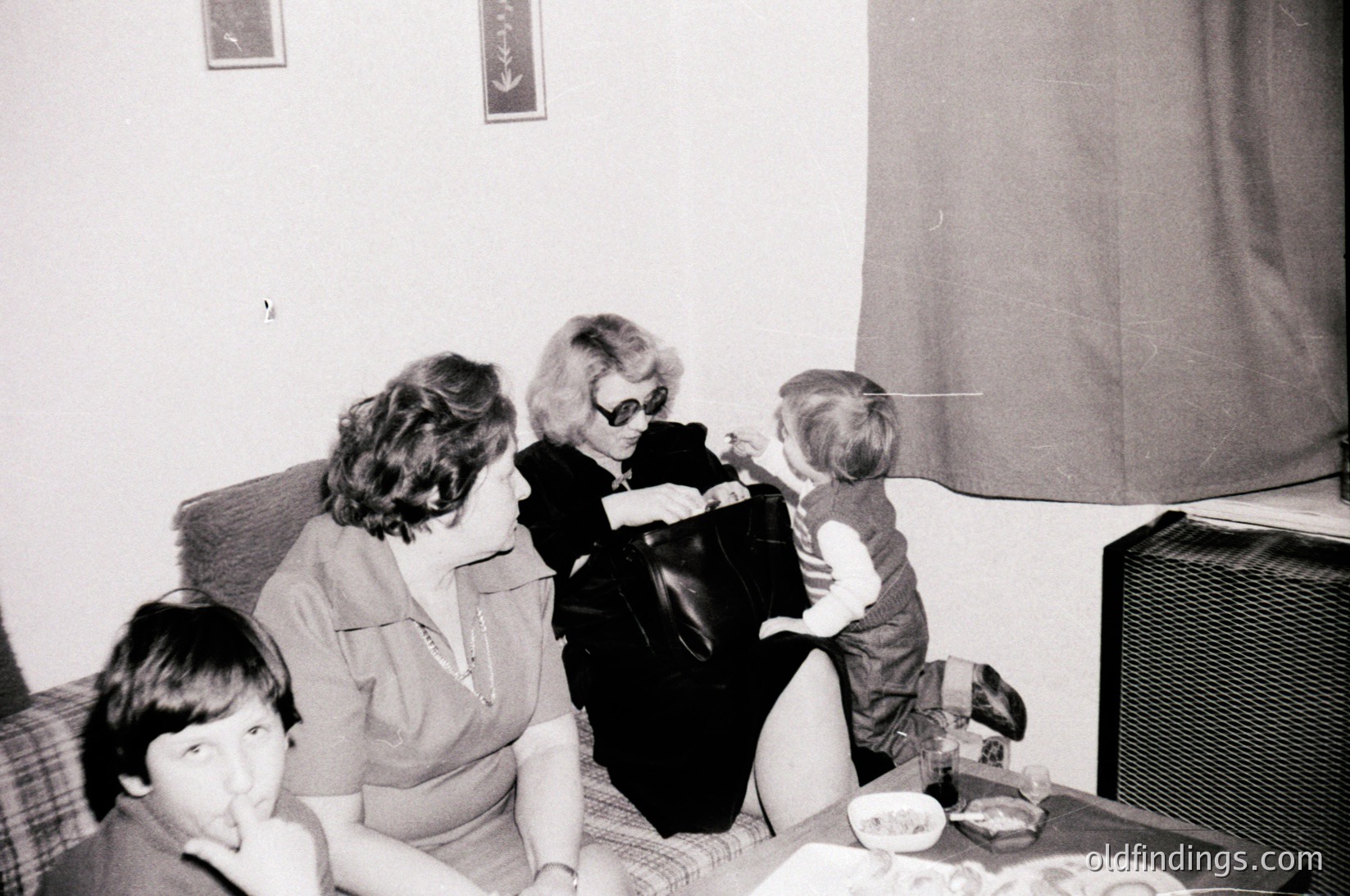Black-and-white snapshot of an indoor family moment, likely mid-20th century. Four individuals—two adults and two children—gather around a low table in a modest living room. The woman in sunglasses adjusts a child’s clothing, while another adult and child sit nearby. A vintage speaker and simple decor (checkered tablecloth, framed art) suggest a mid-century home setting.