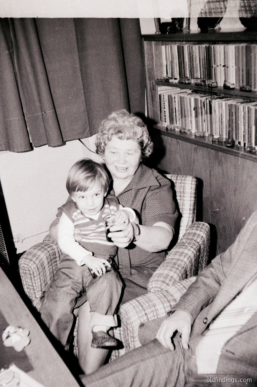 A mid-20th-century indoor portrait showing a woman holding a young child on her lap in a cozy living room. She wears a patterned blouse and skirt, while the child dons a striped shirt and sandals. Behind them, a bookshelf filled with vinyl records and books suggests a mid-century home. The warm lighting and vintage styling evoke 1950s–1960s domestic life.