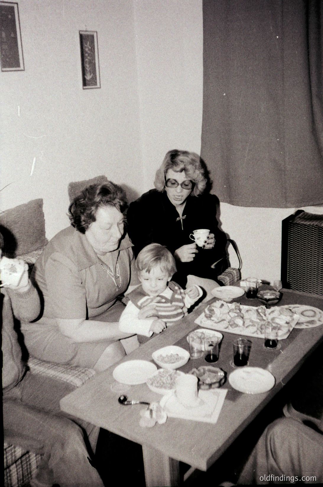 Mid-20th century indoor family gathering at a wooden table with plates of food, including pastries and a cake. Three women and a young child seated; one woman holds a teacup. Plain curtains and a speaker suggest a modest home setting, likely 1960s–1970s.