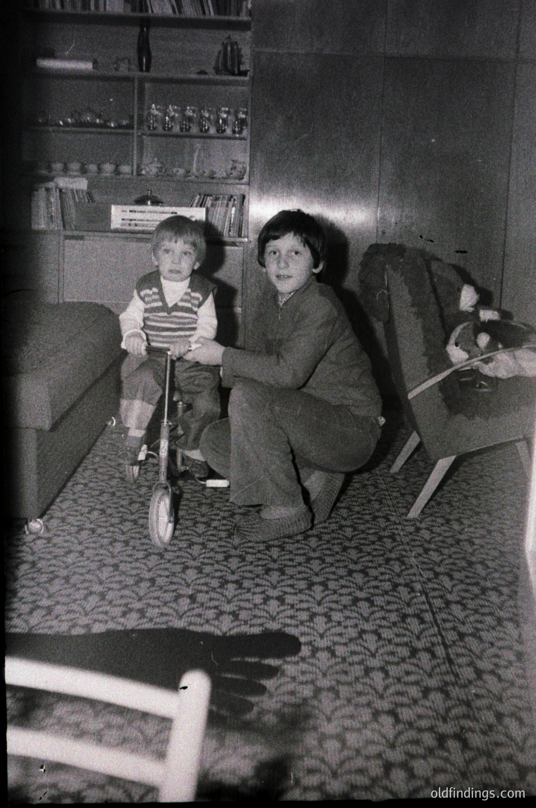 Mid-20th century indoor scene: two children in a dimly lit living room, one seated on a balance bike, the other kneeling beside. Bookshelves with glass-fronted cabinets and patterned carpet.